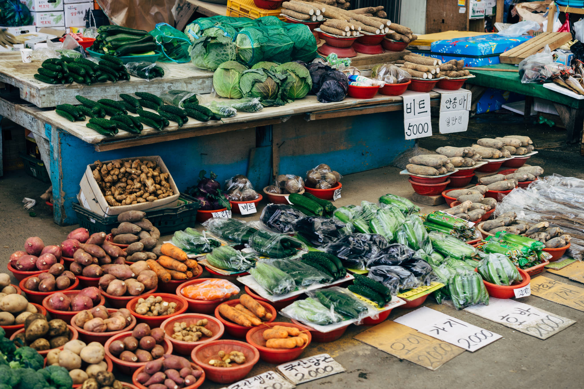 Busan market vegetable stall with various produce including cucumbers, ginger, potatoes, carrots, and other vegetables in red bowls.