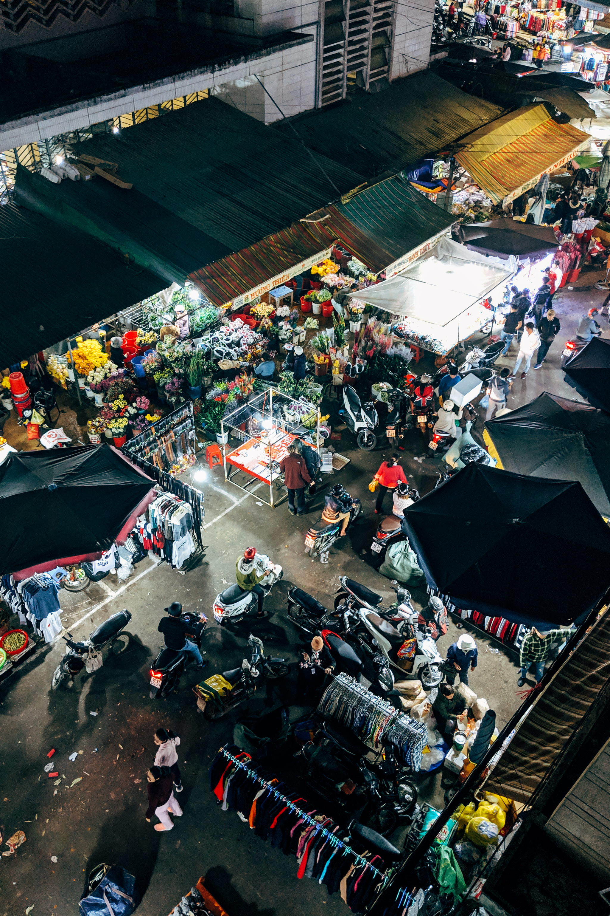 Aerial view of a bustling night market with vendors selling flowers, clothing, and other goods; many motorbikes are parked amongst the stalls.