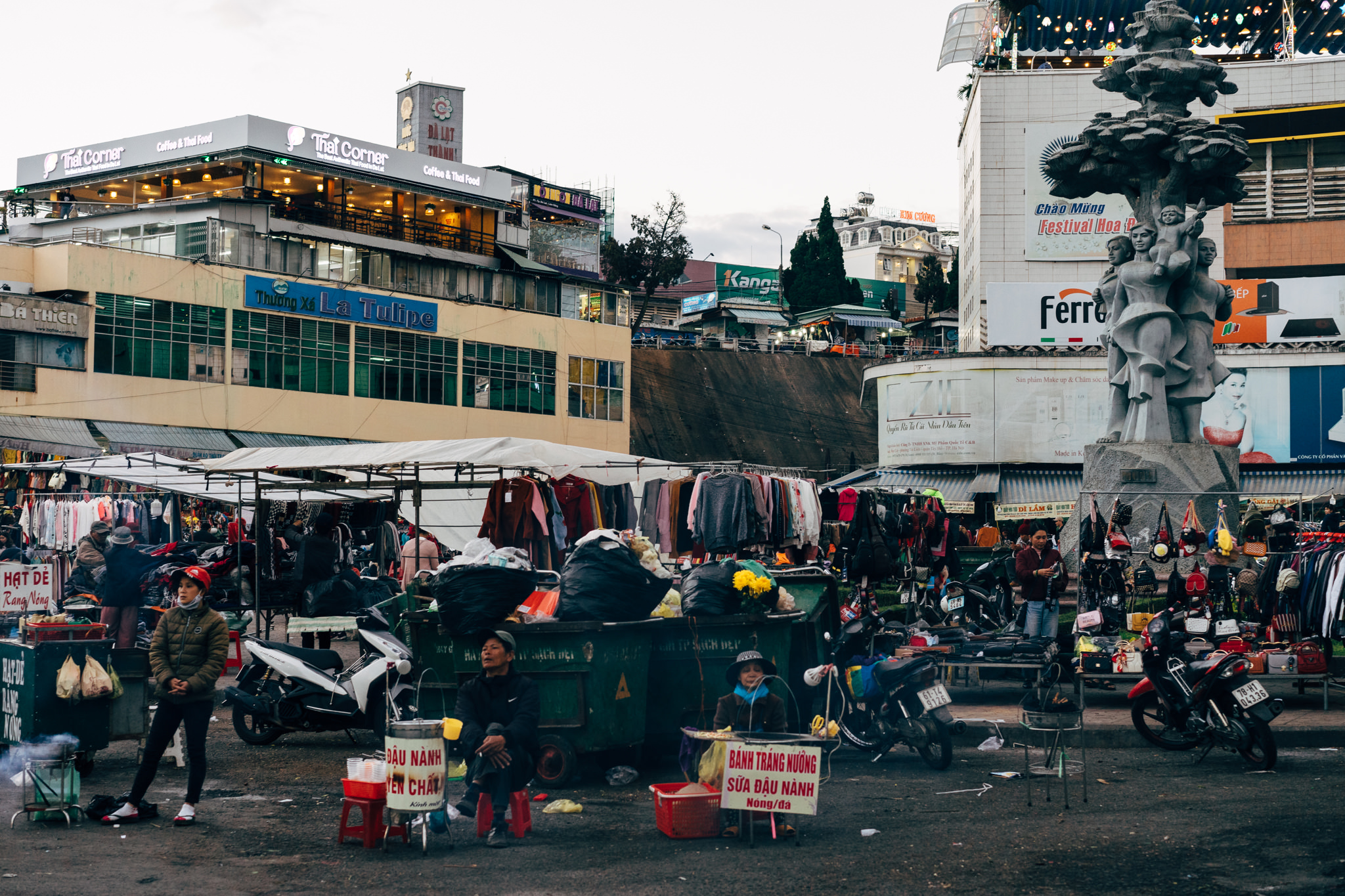 Da Lat market scene with vendors and shoppers near a statue.