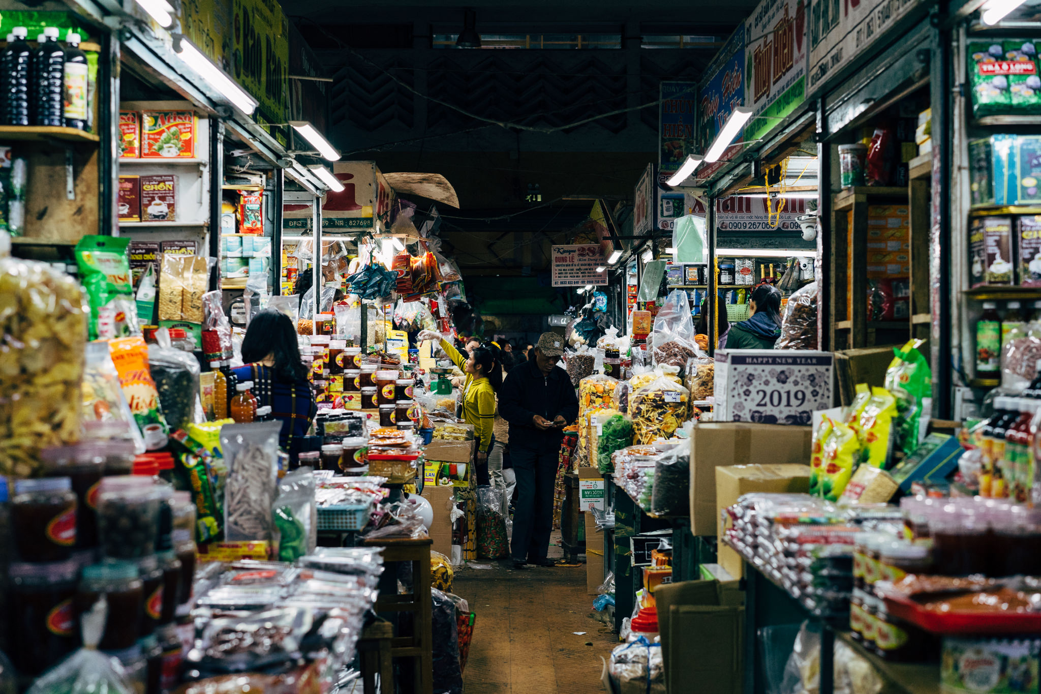 A bustling indoor market in Da Lat, Vietnam, overflowing with packaged goods and snacks.