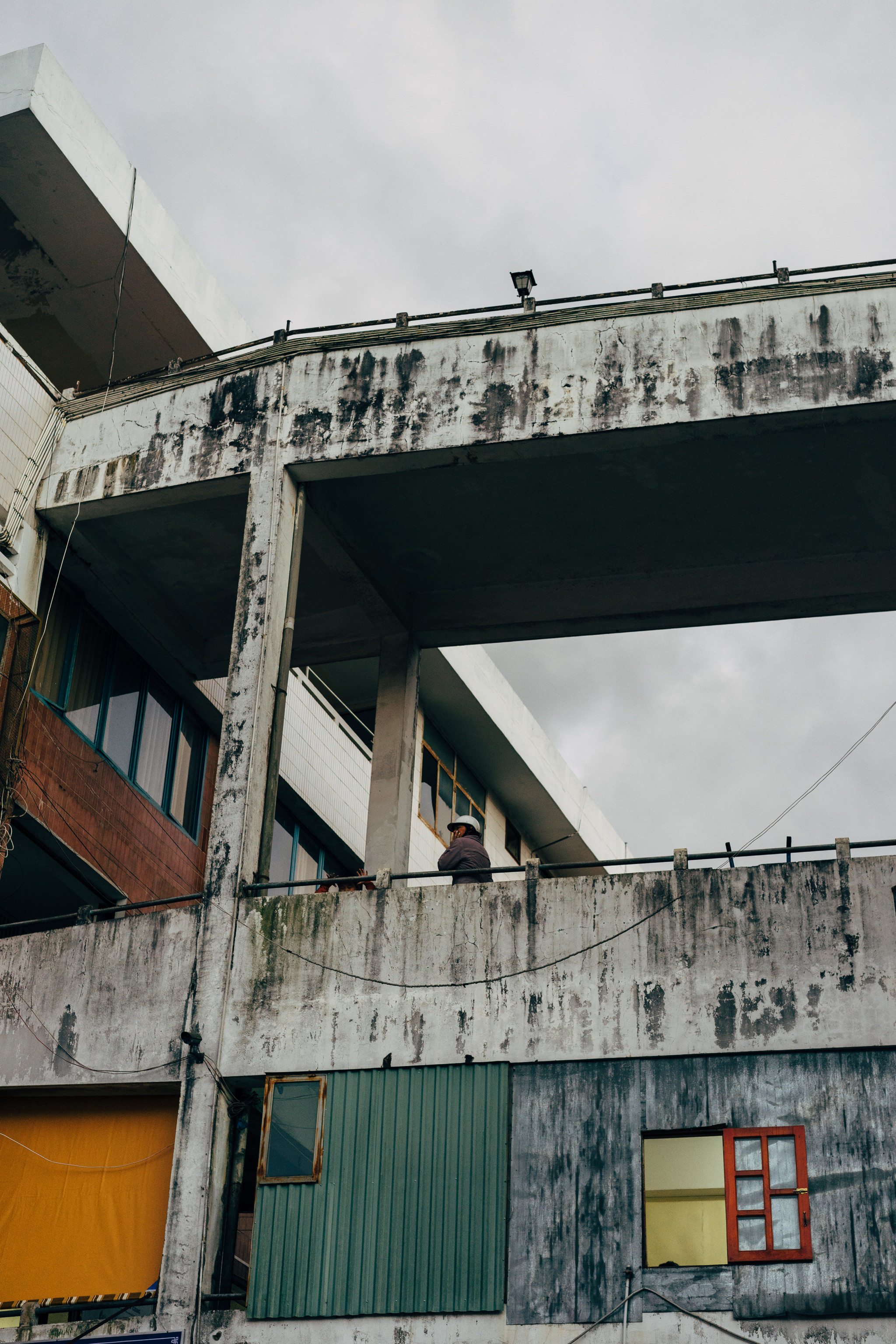 A person stands on a weathered concrete balcony overlooking a dilapidated building.