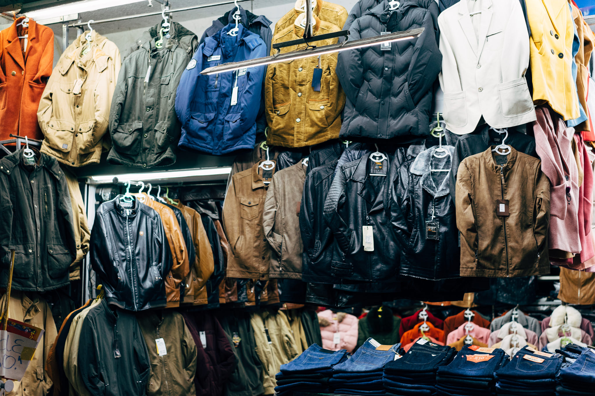 Various jackets and coats hanging on racks in a market, with jeans stacked below.