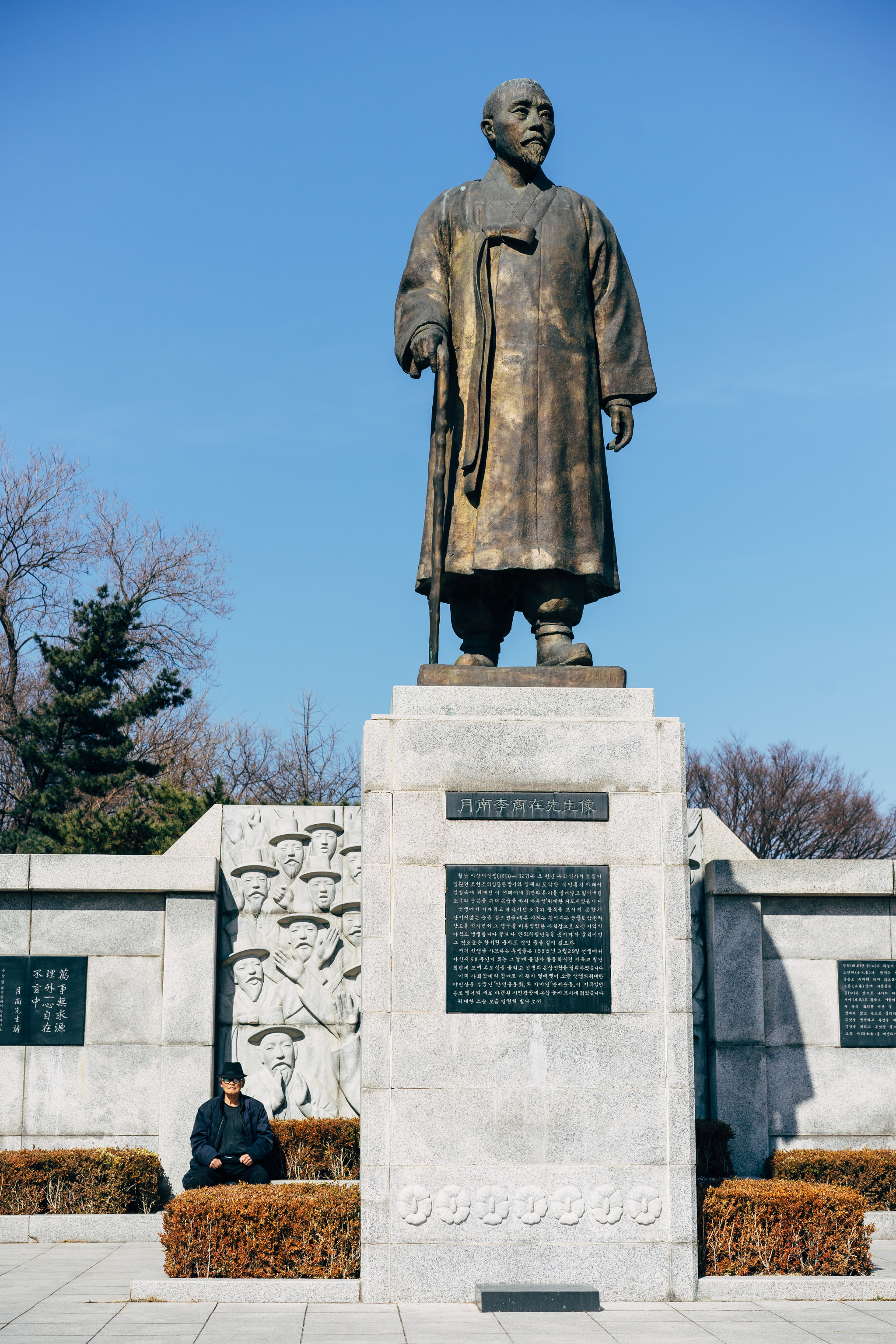 Large bronze statue of a man in traditional Korean clothing holding a staff, situated on a stone monument with Korean text.
