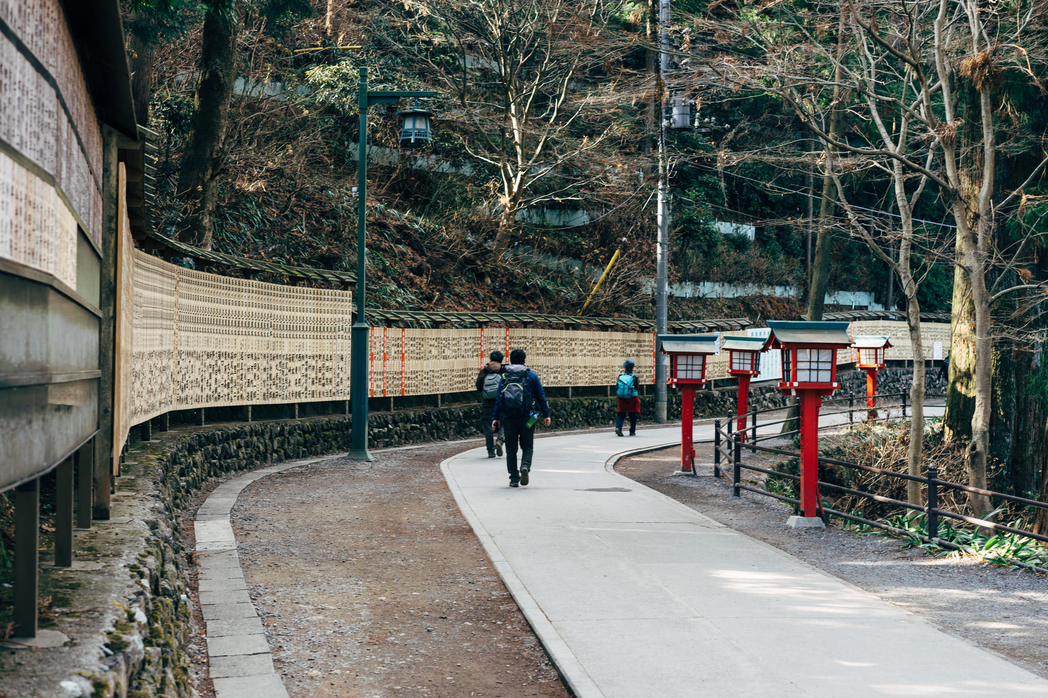 Three people walk away from the camera on a paved path lined with a wall of inscribed tablets and red lanterns.