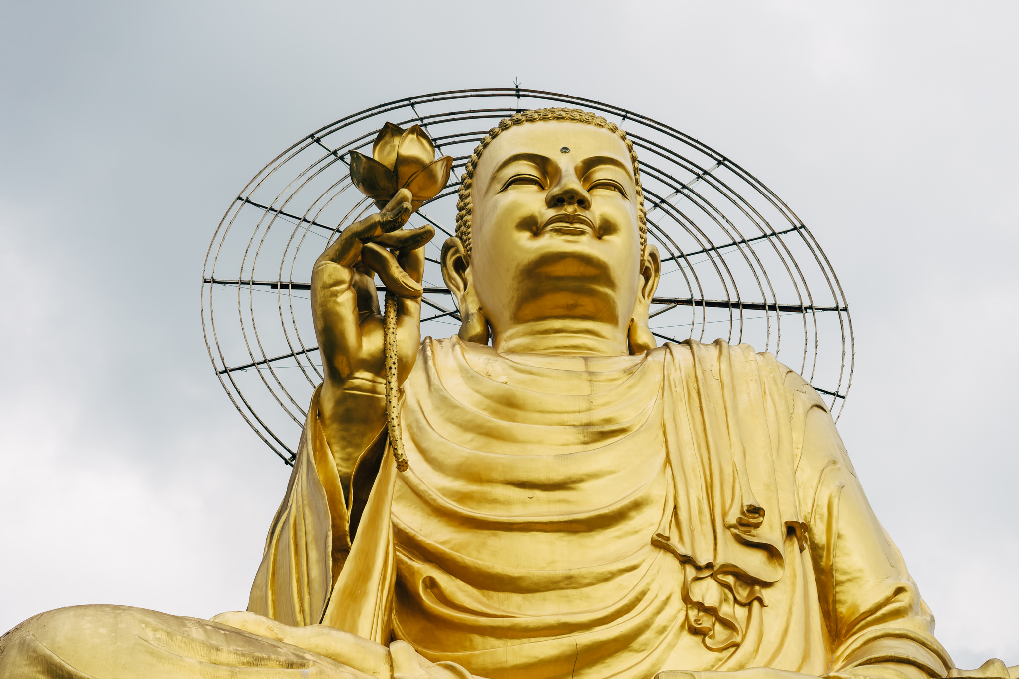 Large golden Buddha statue holding a lotus flower, with a circular metal structure behind it.