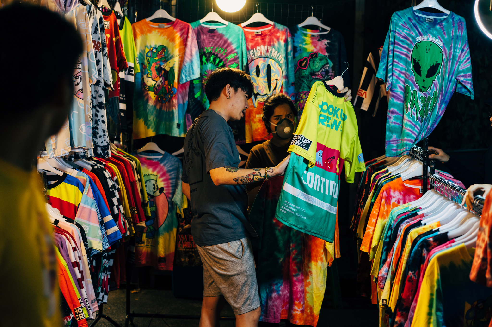 Two people shopping for colorful tie-dye shirts at a Bangkok market.