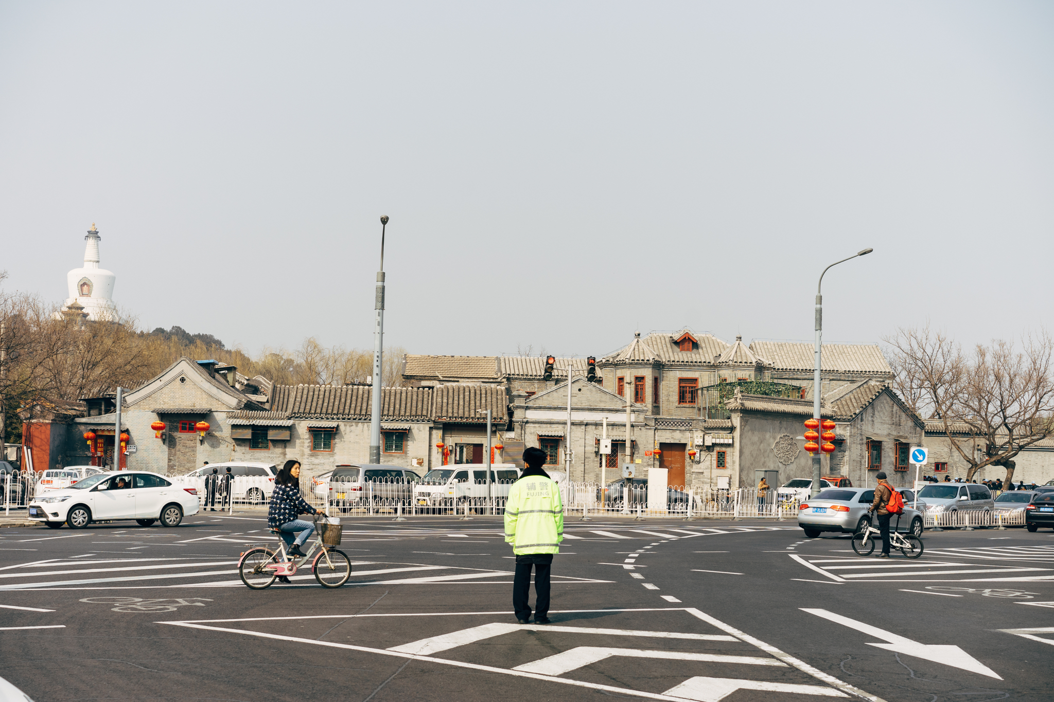 Beijing street scene: police officer directing traffic at an intersection with cyclists and cars; traditional buildings and a white pagoda in the background.