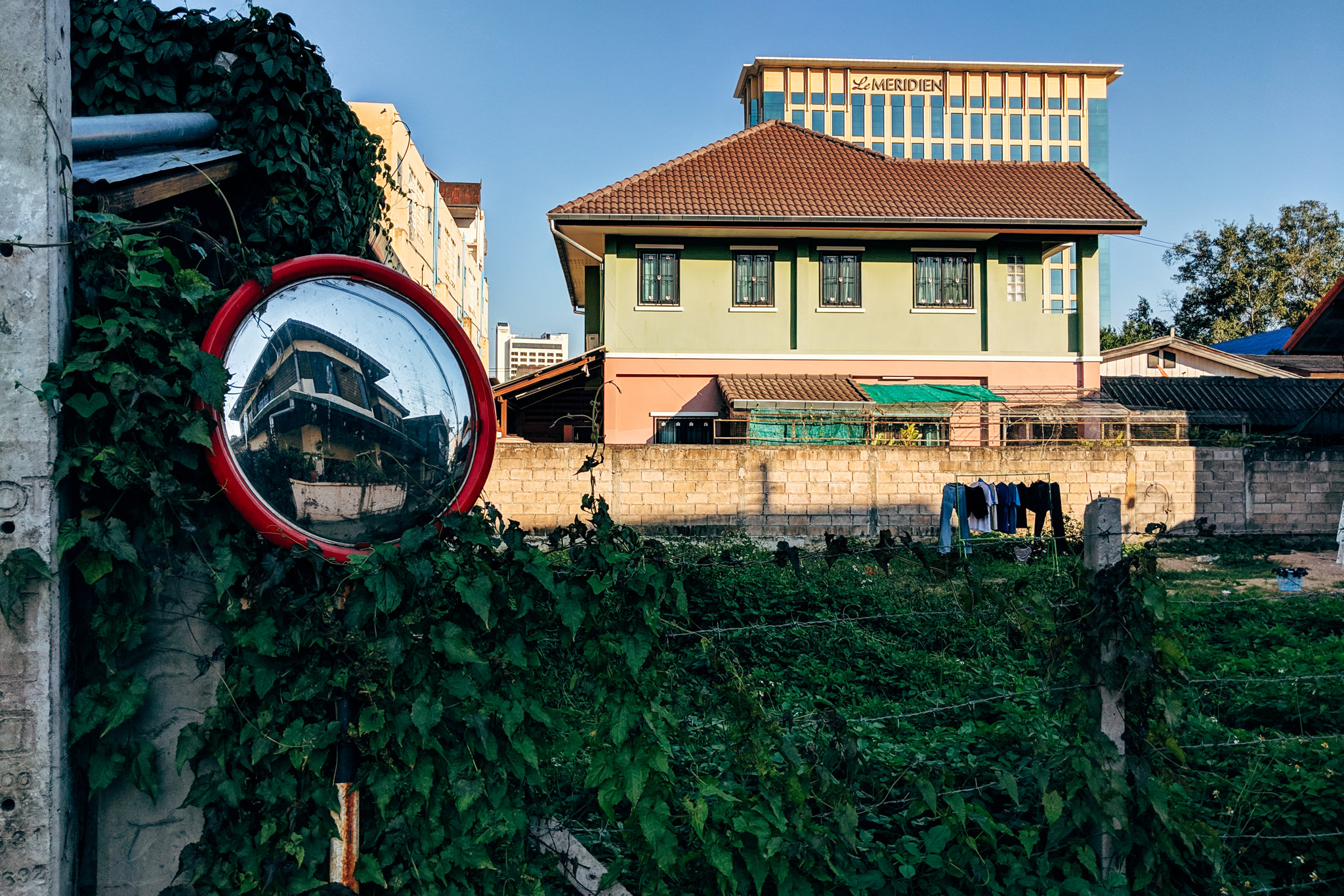 A red convex mirror reflecting a house, partially obscured by foliage, with a green house and Le Meridien hotel in the background.