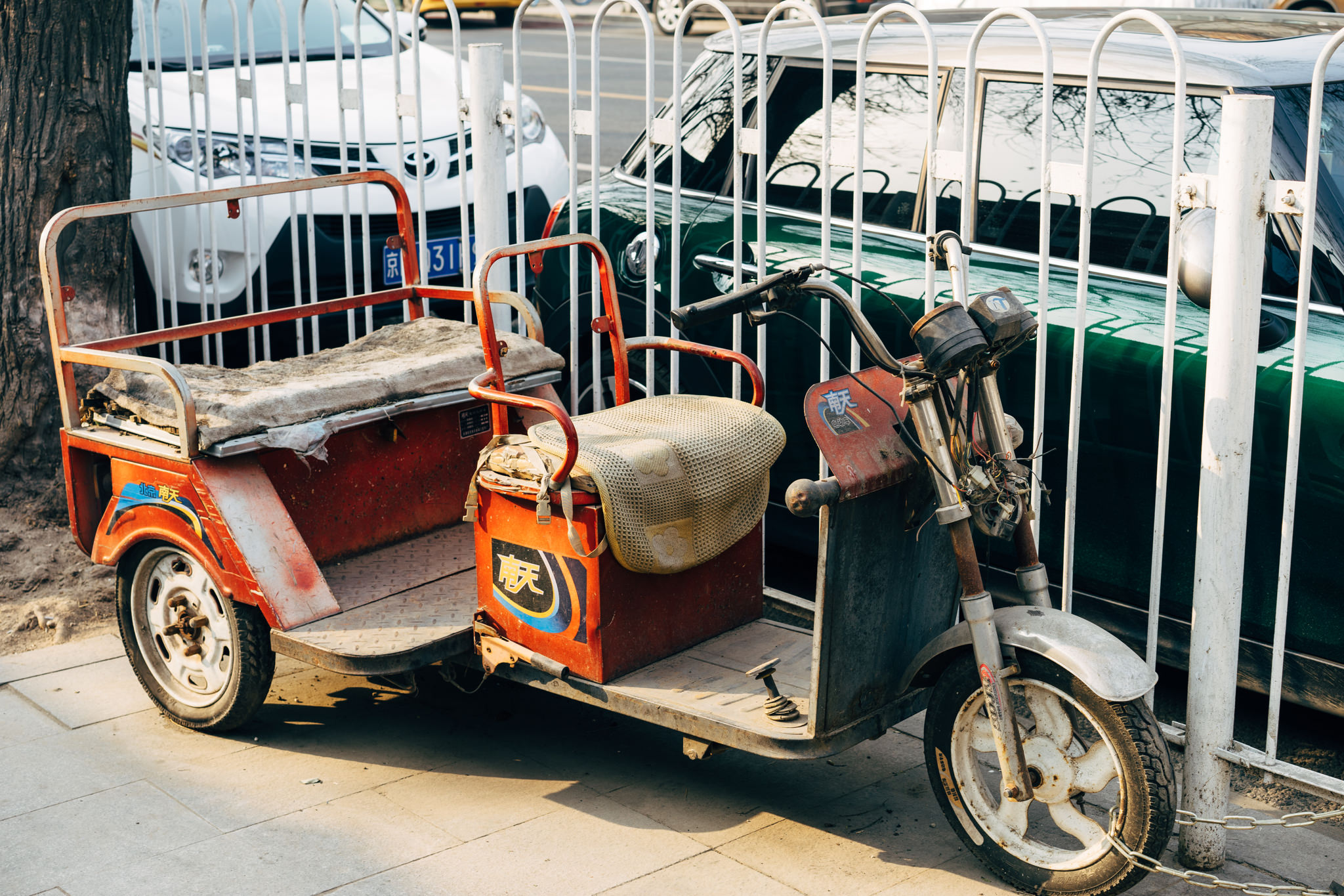 Worn red electric tricycle parked on city street, chained to fence behind parked cars.