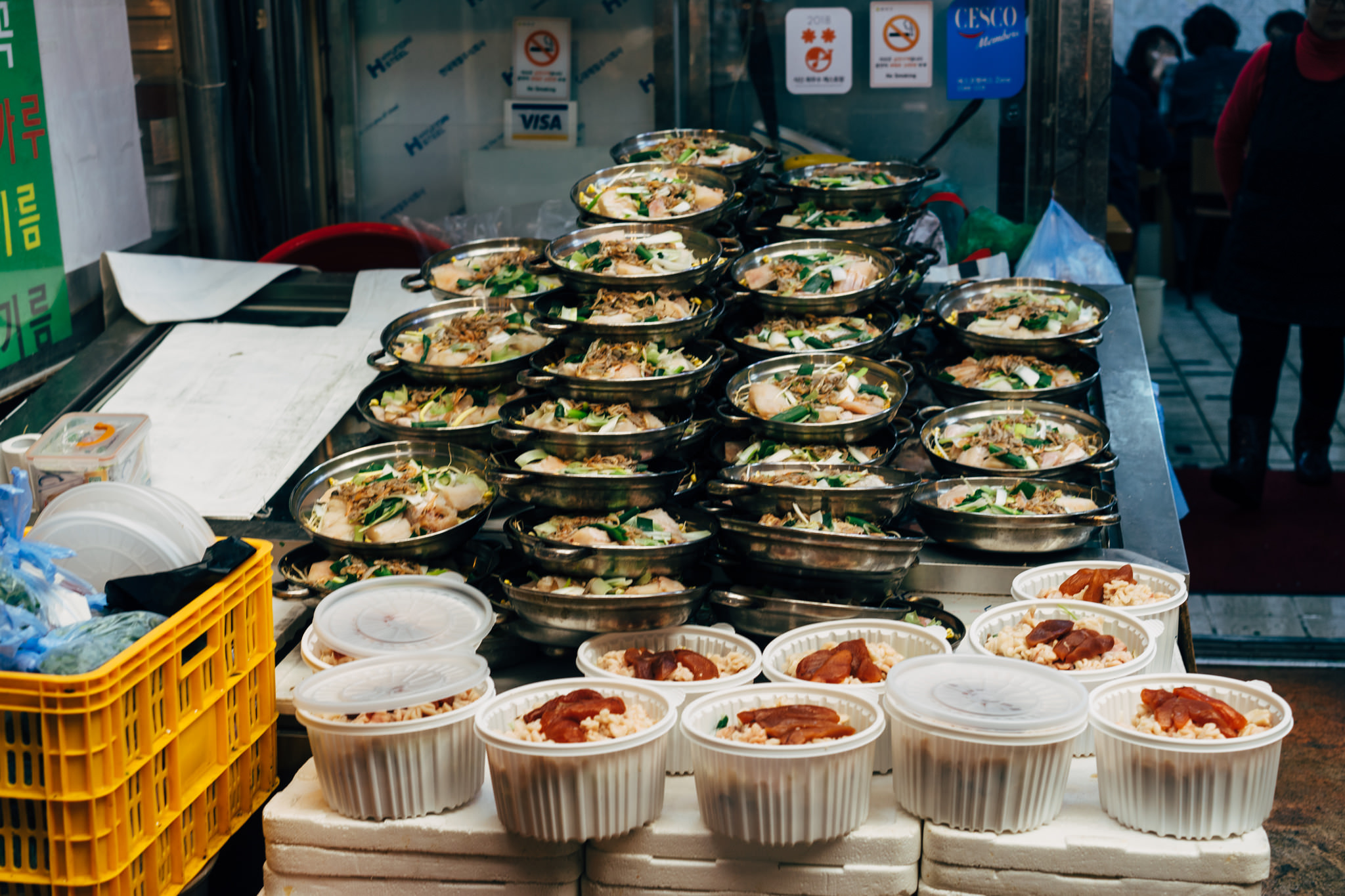 Korean market food stall with numerous metal pans of food and containers of rice dishes.