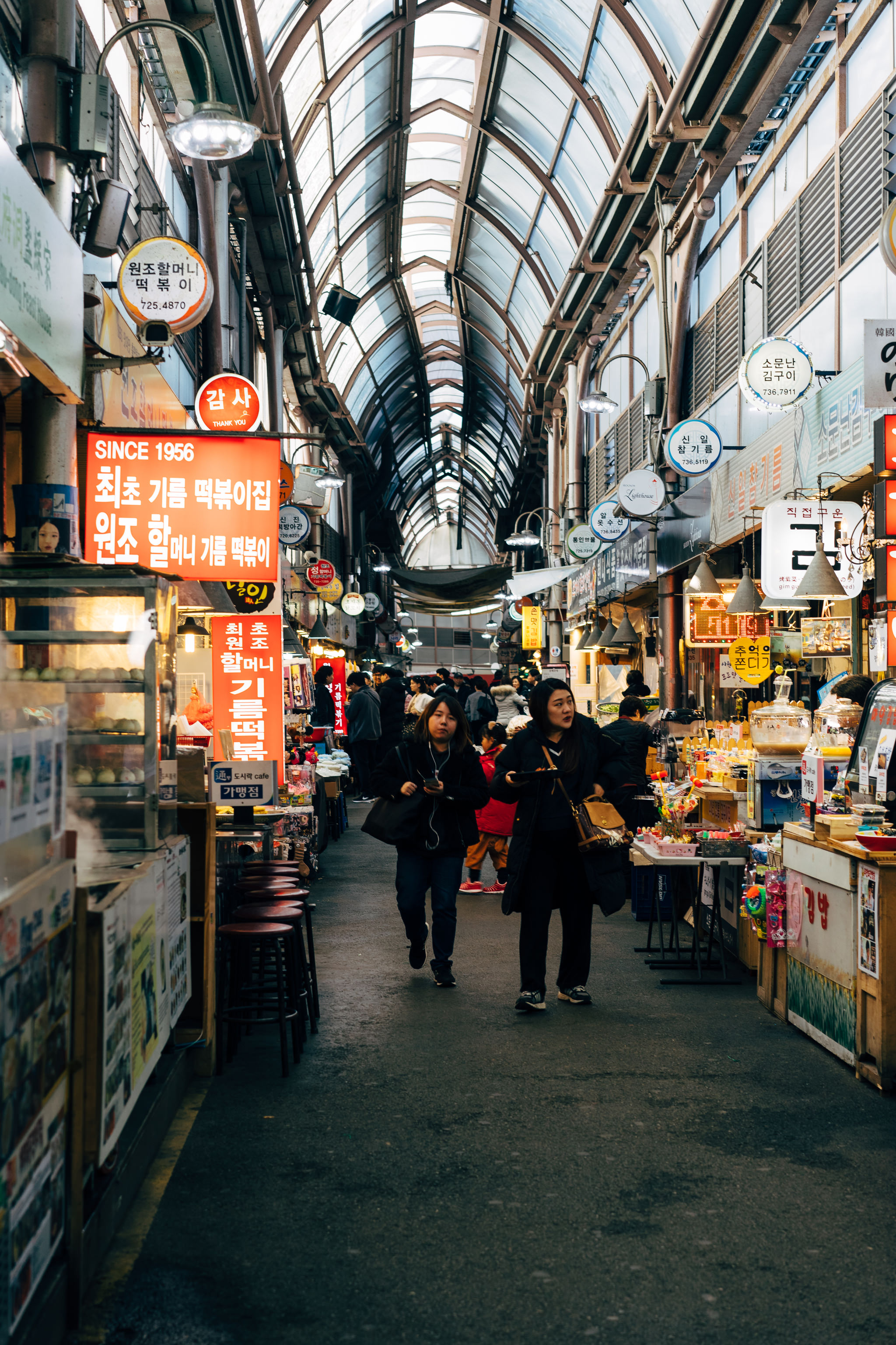 Two women walking through a bustling Korean market with numerous food stalls and signage.
