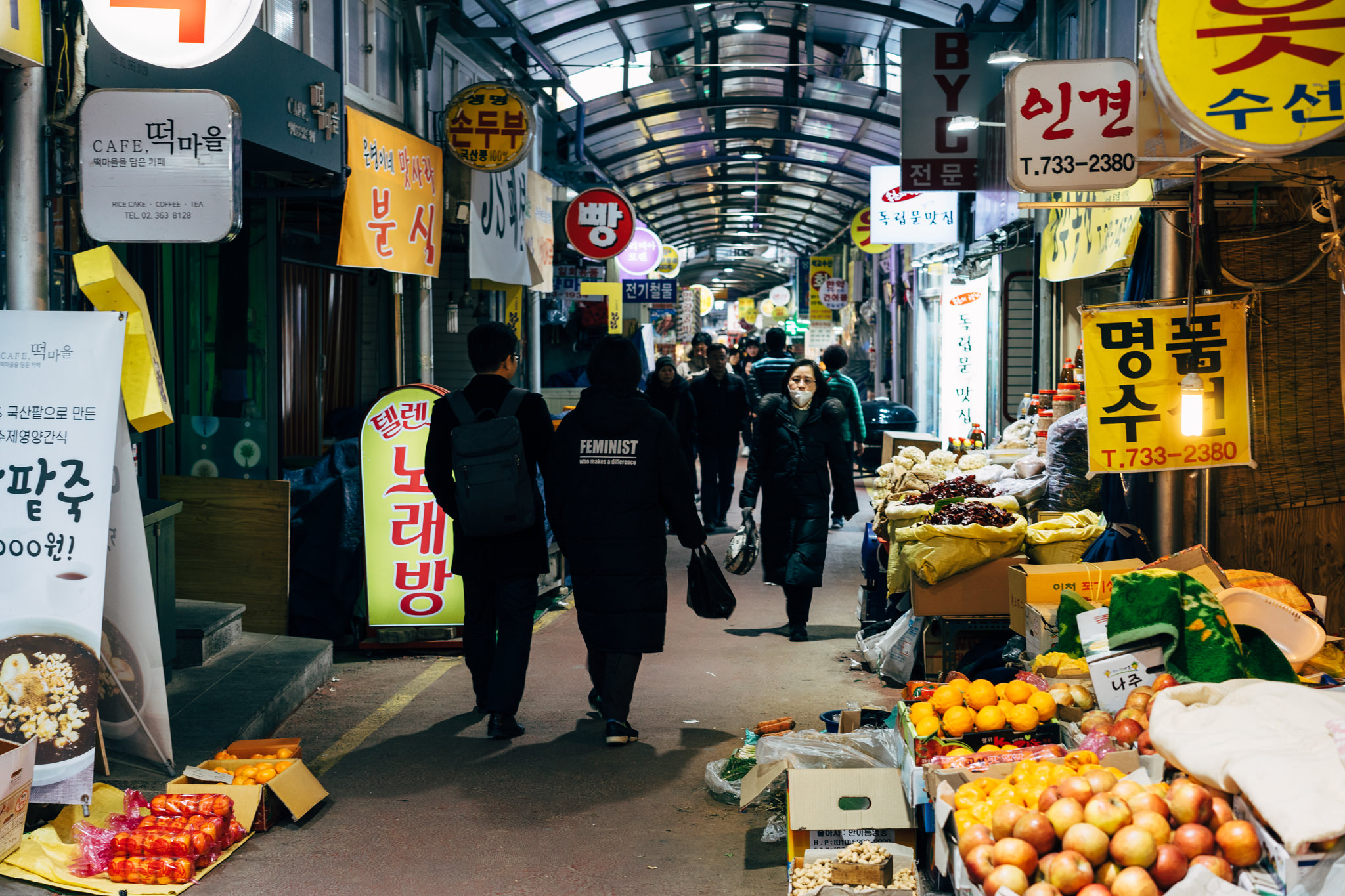 People walk through a narrow, covered market in Korea, lined with shops and produce.