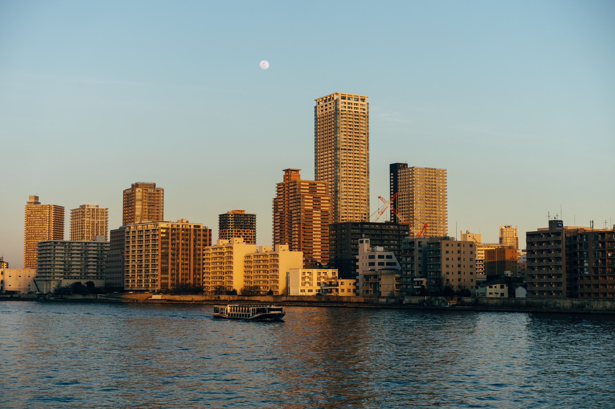 Tokyo cityscape at sunset with a boat on the river and a full moon in the sky.