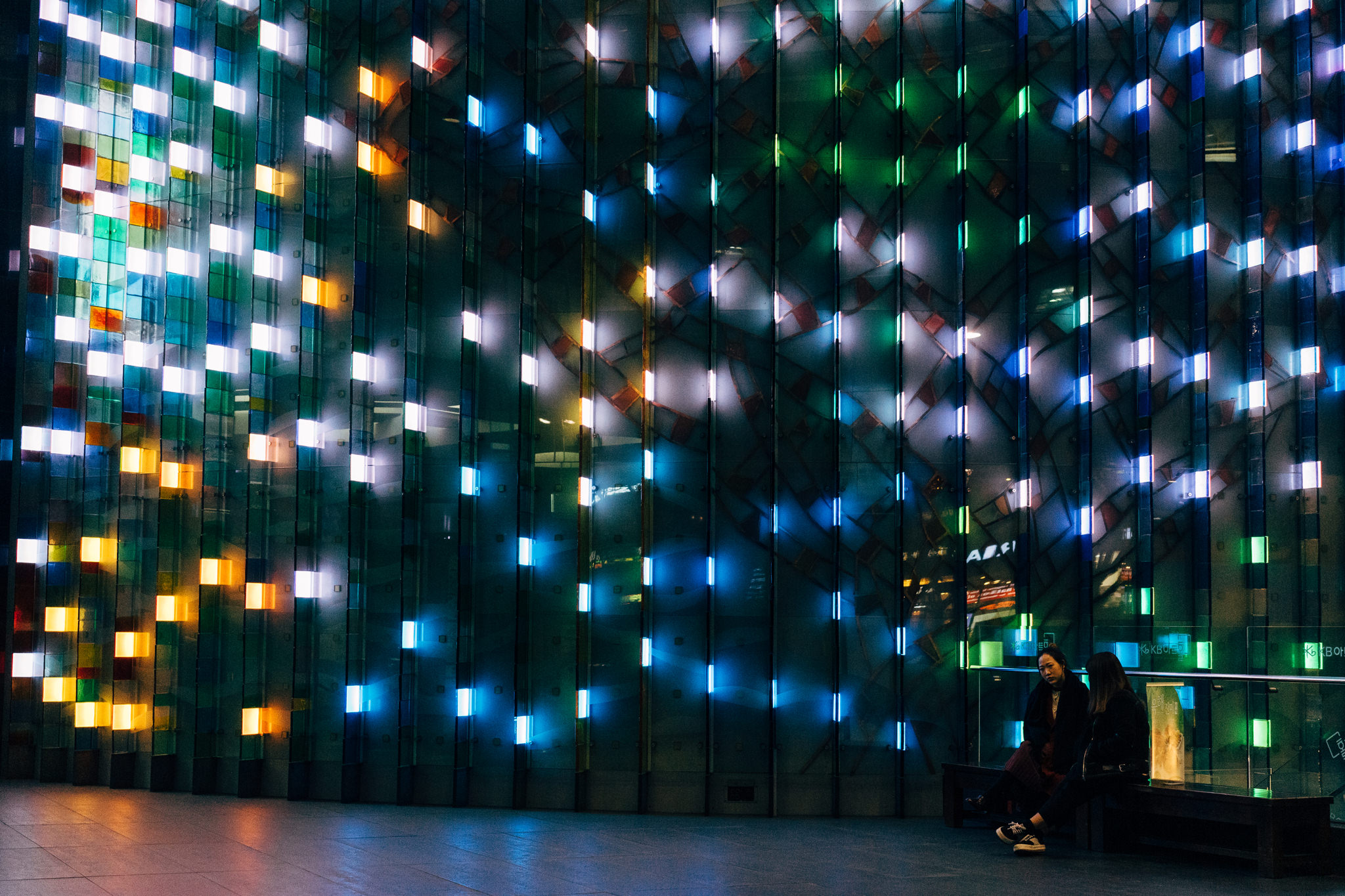Two people sitting on a bench in front of a colorful, illuminated glass wall at night.