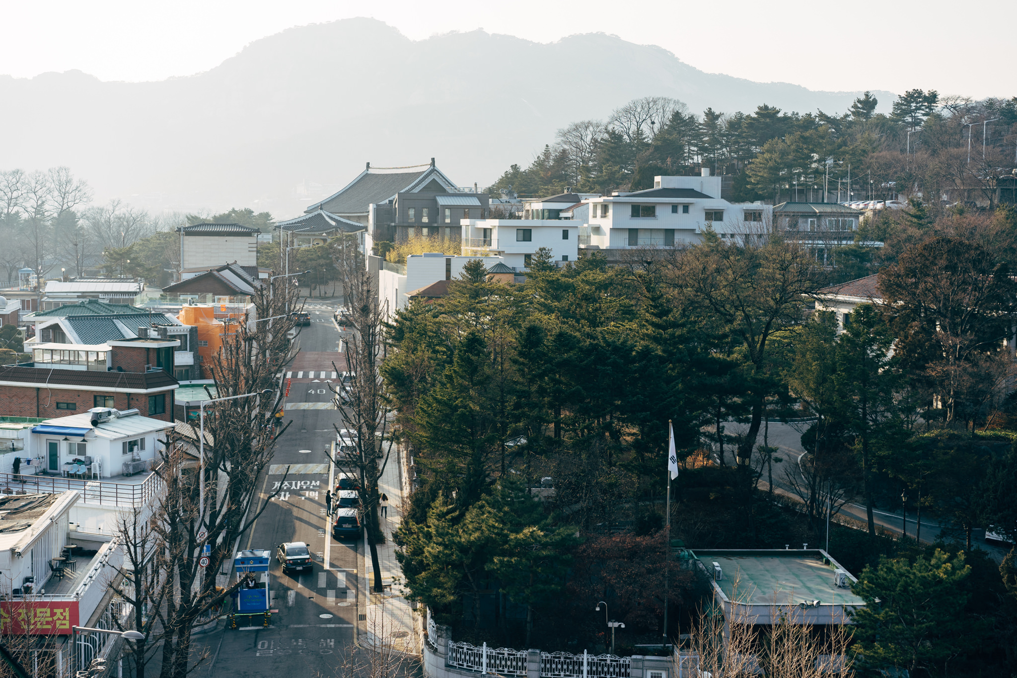 Aerial view of a Seoul street with traditional and modern buildings, trees, and mountains in the background.