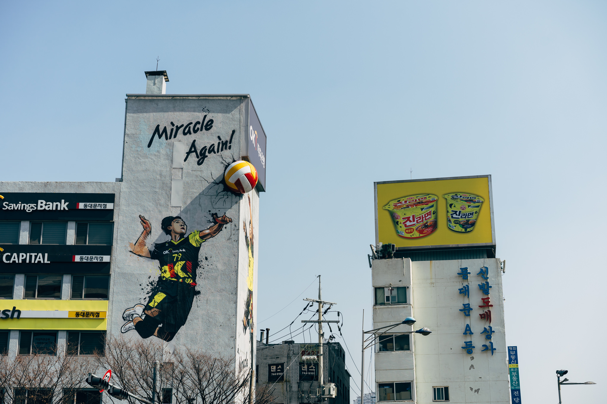 Seoul street scene with a mural of a volleyball player, 'Miracle Again!' text, and building-mounted advertisements.