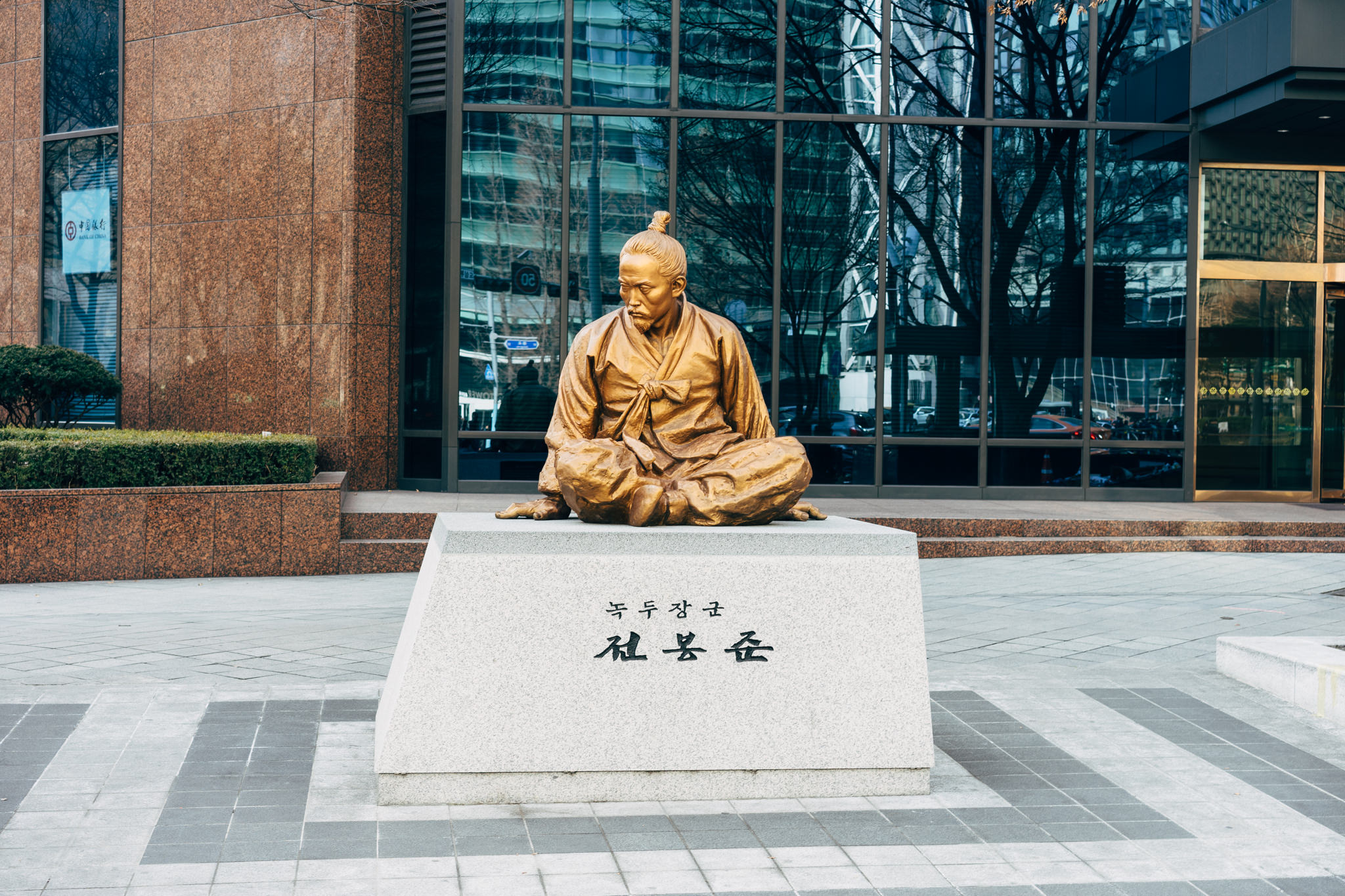 Bronze statue of a seated man in traditional Korean clothing outside a modern building in Seoul, South Korea.