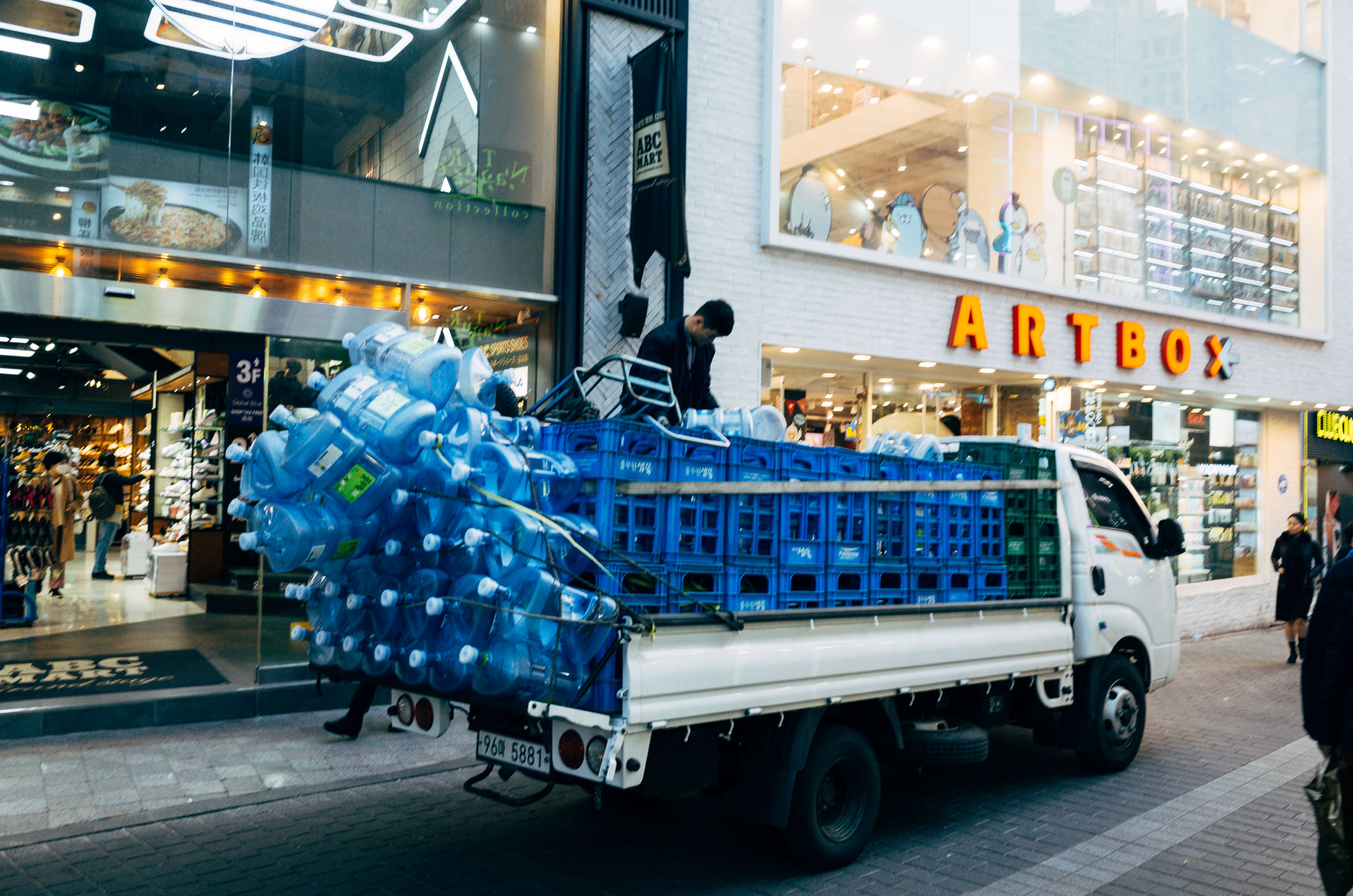 A white truck loaded with large water bottles in blue crates parked on a city street in Seoul, South Korea.