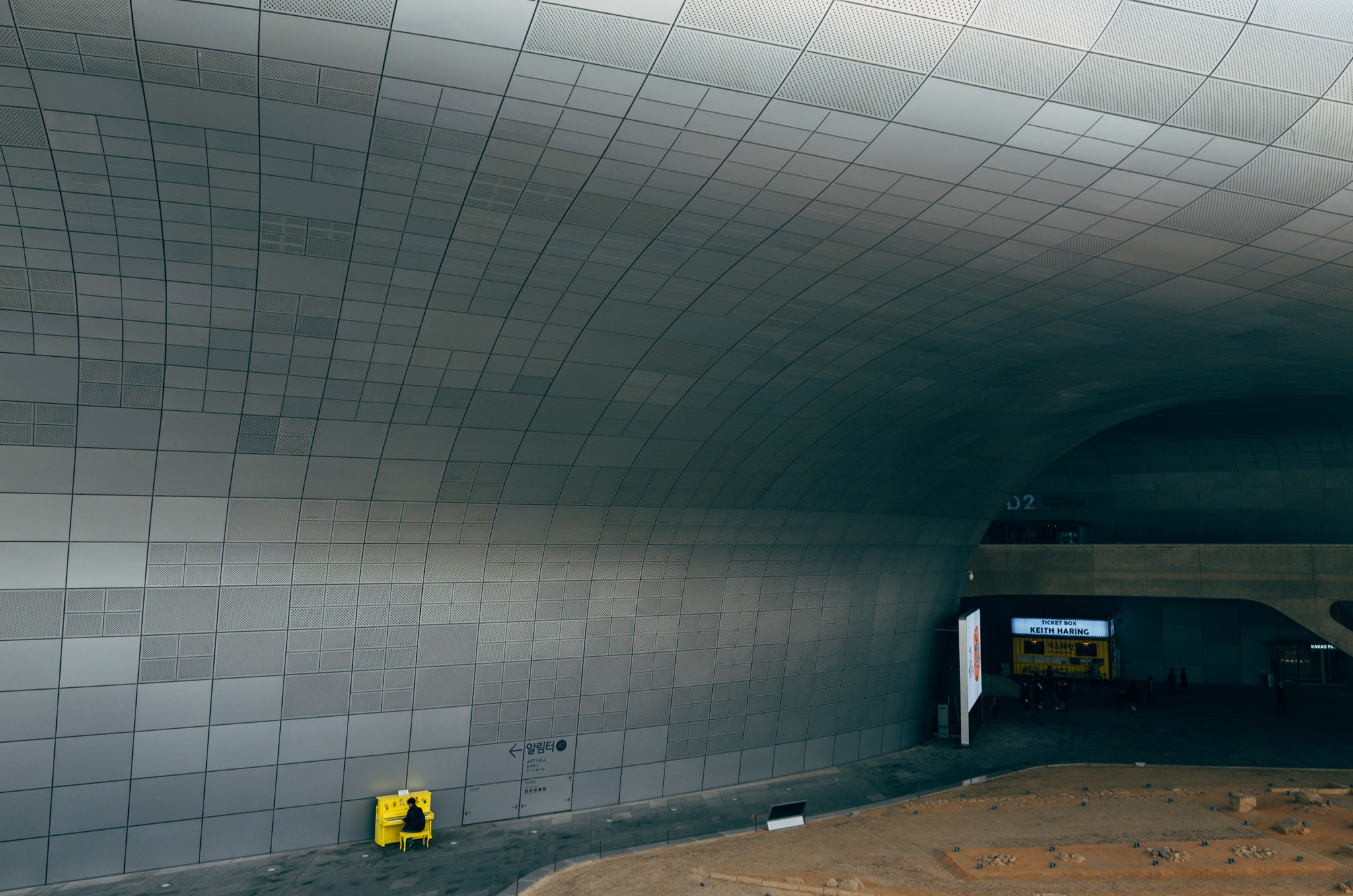 Person playing a yellow piano against a large, curved gray wall.