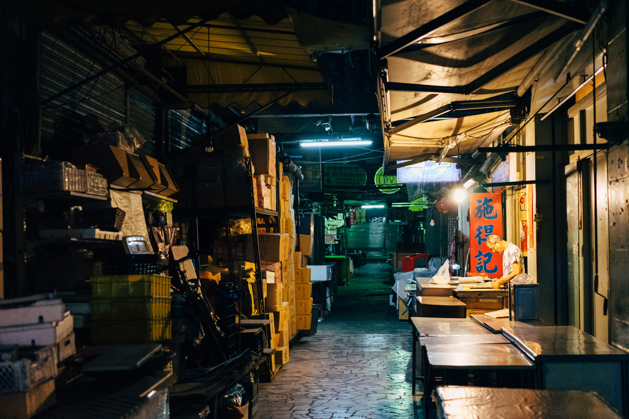 A person works at a food stall in a dimly lit, cluttered alleyway at night.