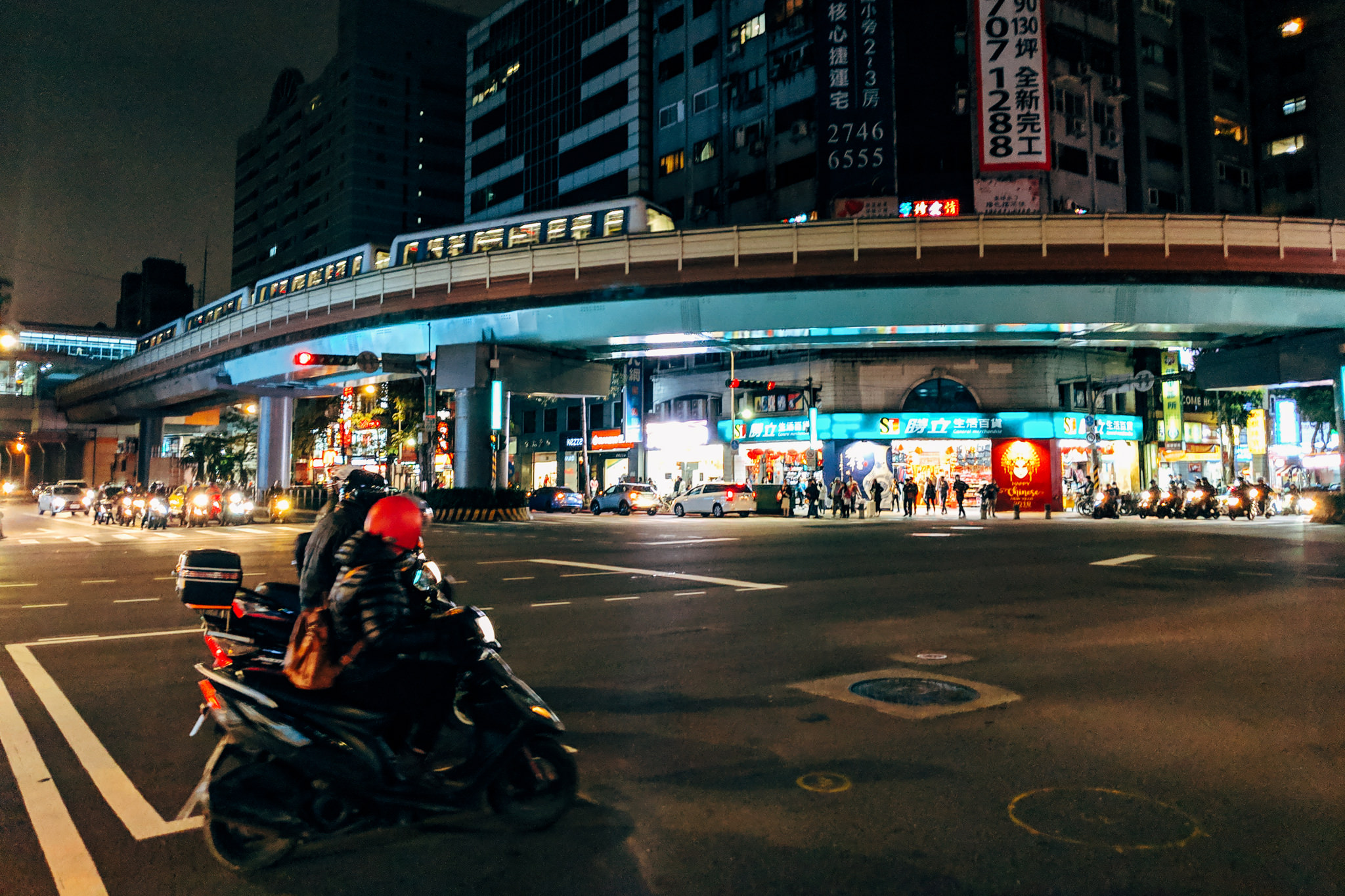 Nighttime street scene in Taipei, Taiwan, showing a motor scooter at an intersection under an elevated train track.