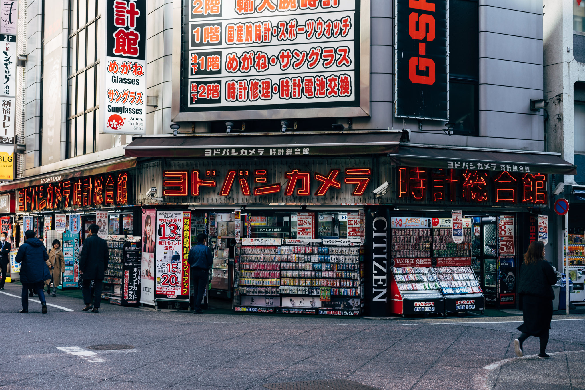 Tokyo street scene with a crowded electronics and watch store, ヨドバシカメラ 時計総合館, featuring numerous signs in Japanese and English.