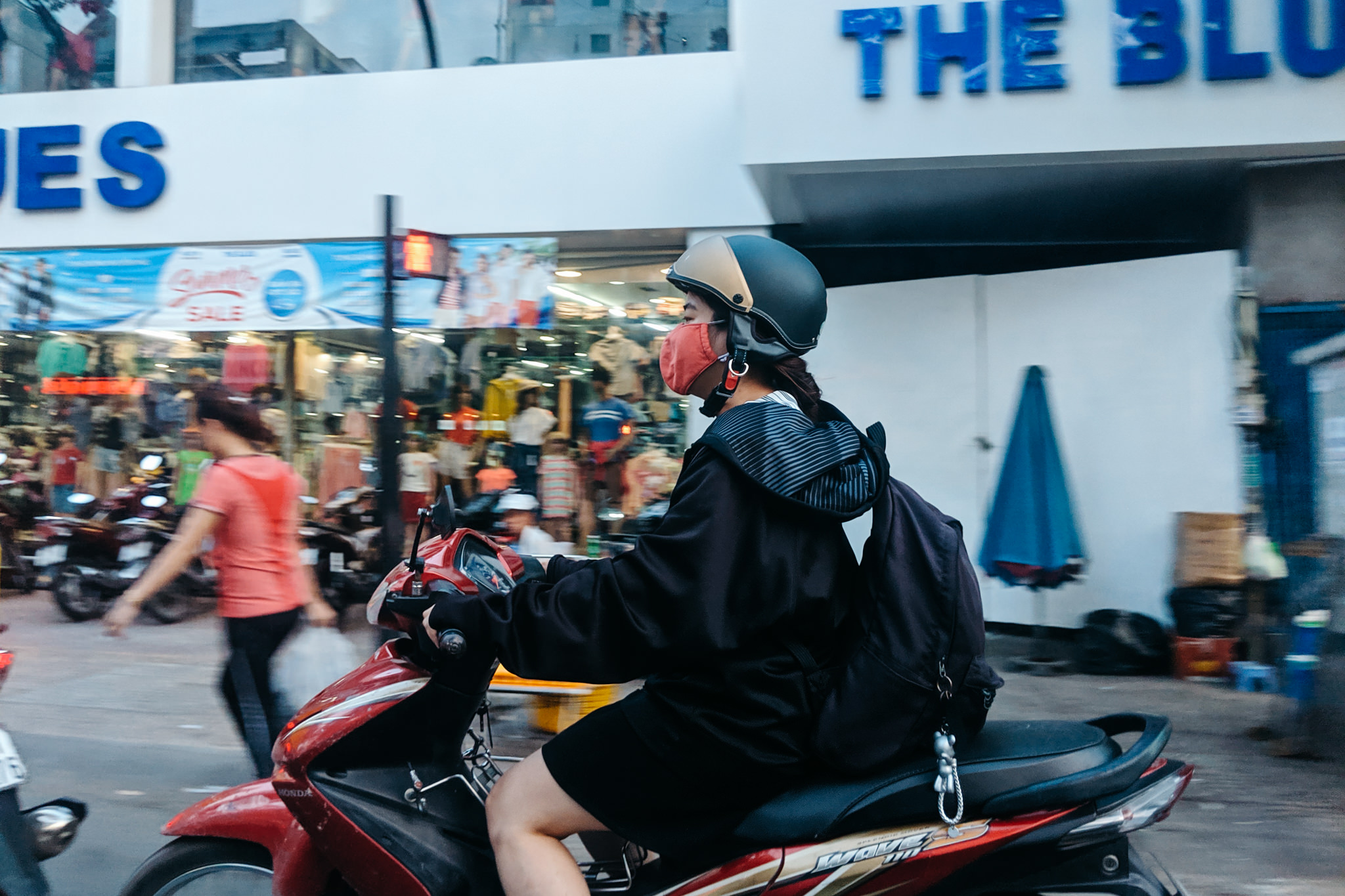 Woman wearing helmet and facemask riding red scooter.