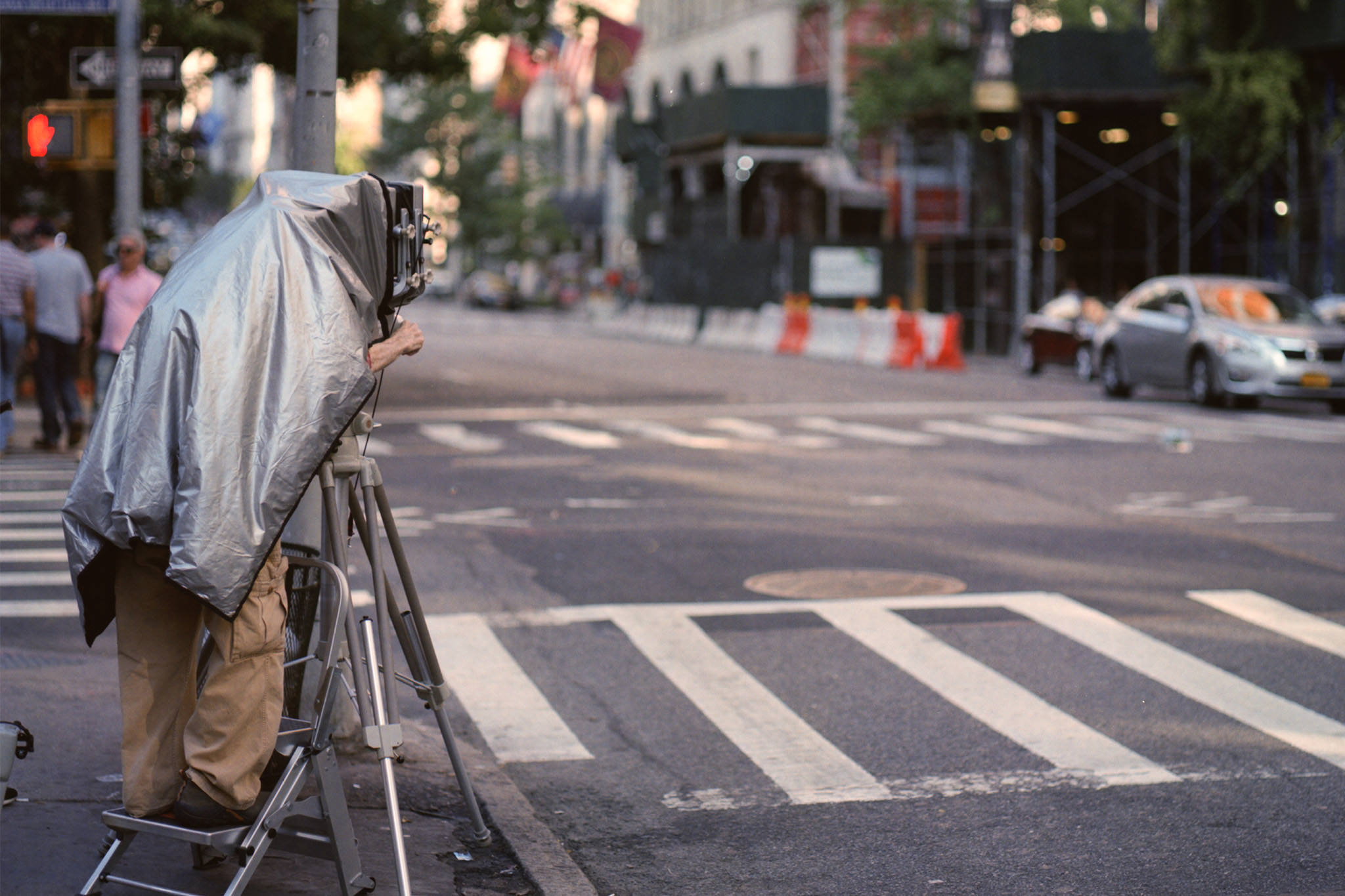 Person using a large vintage camera on a tripod, covered by a silver tarp, taking a photograph on a New York City street.