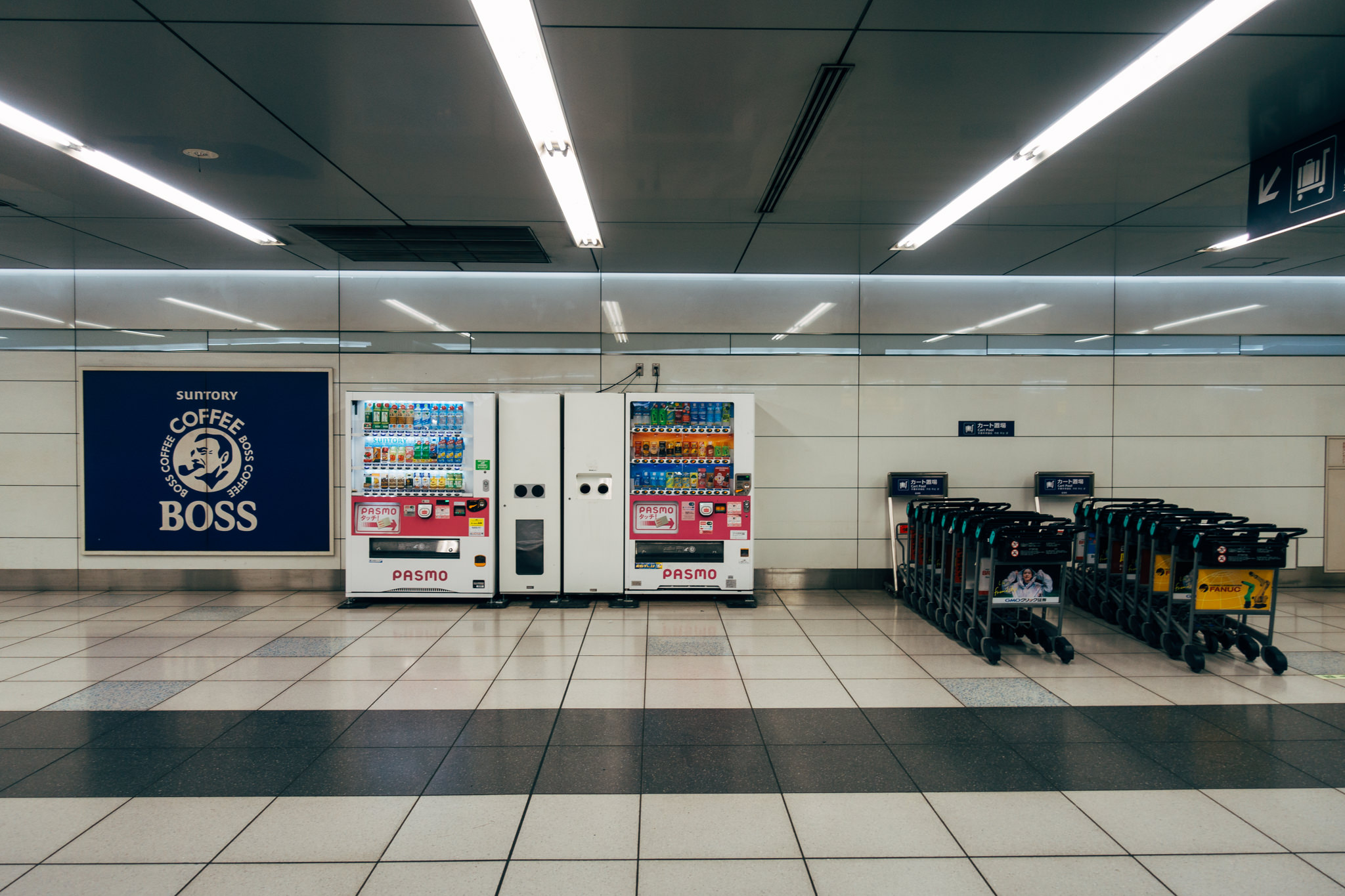 Two beverage vending machines and rows of luggage carts in a Japanese transit station.