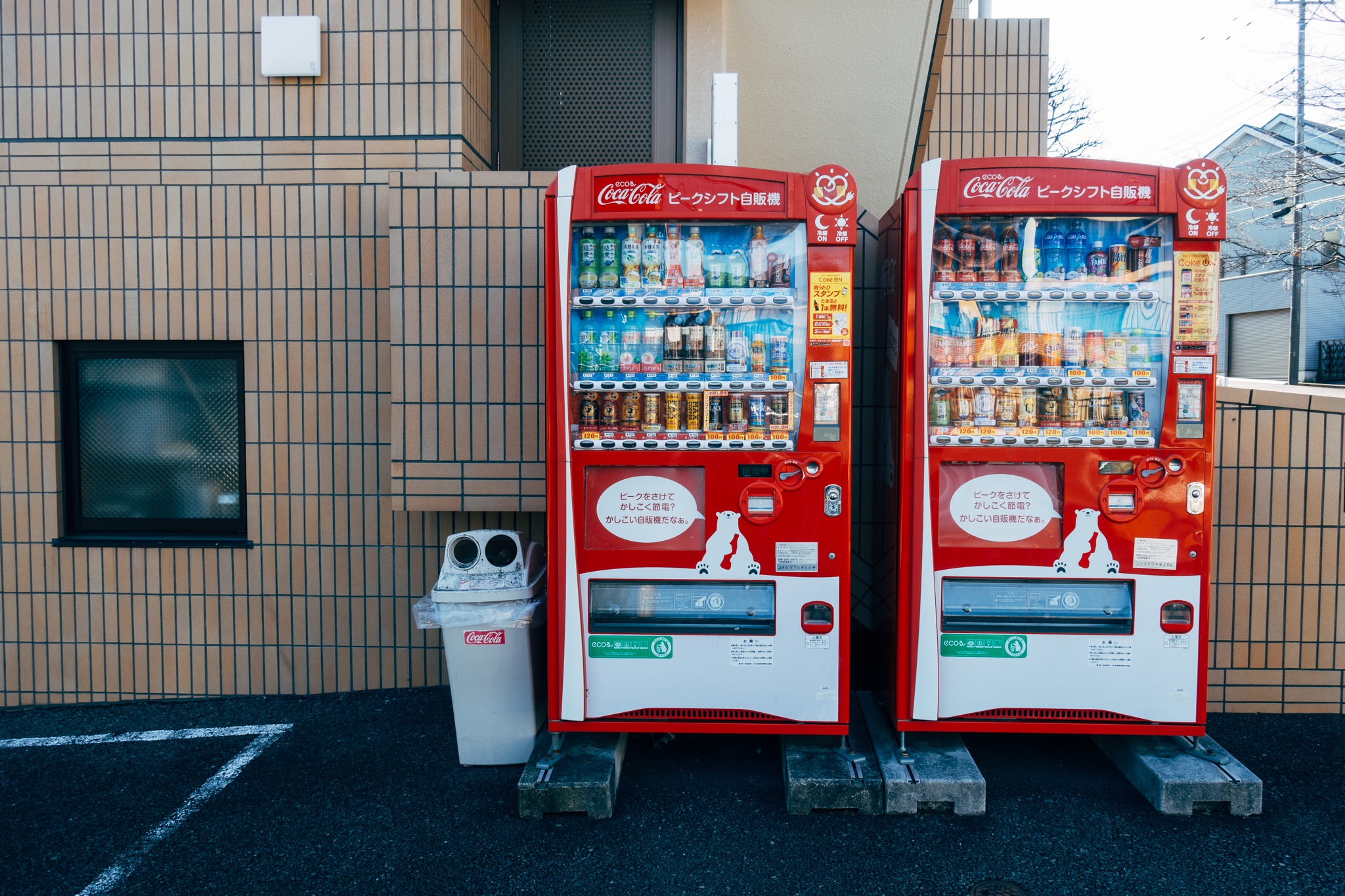 Two red Coca-Cola vending machines in Japan, stocked with beverages, and a small trash can.