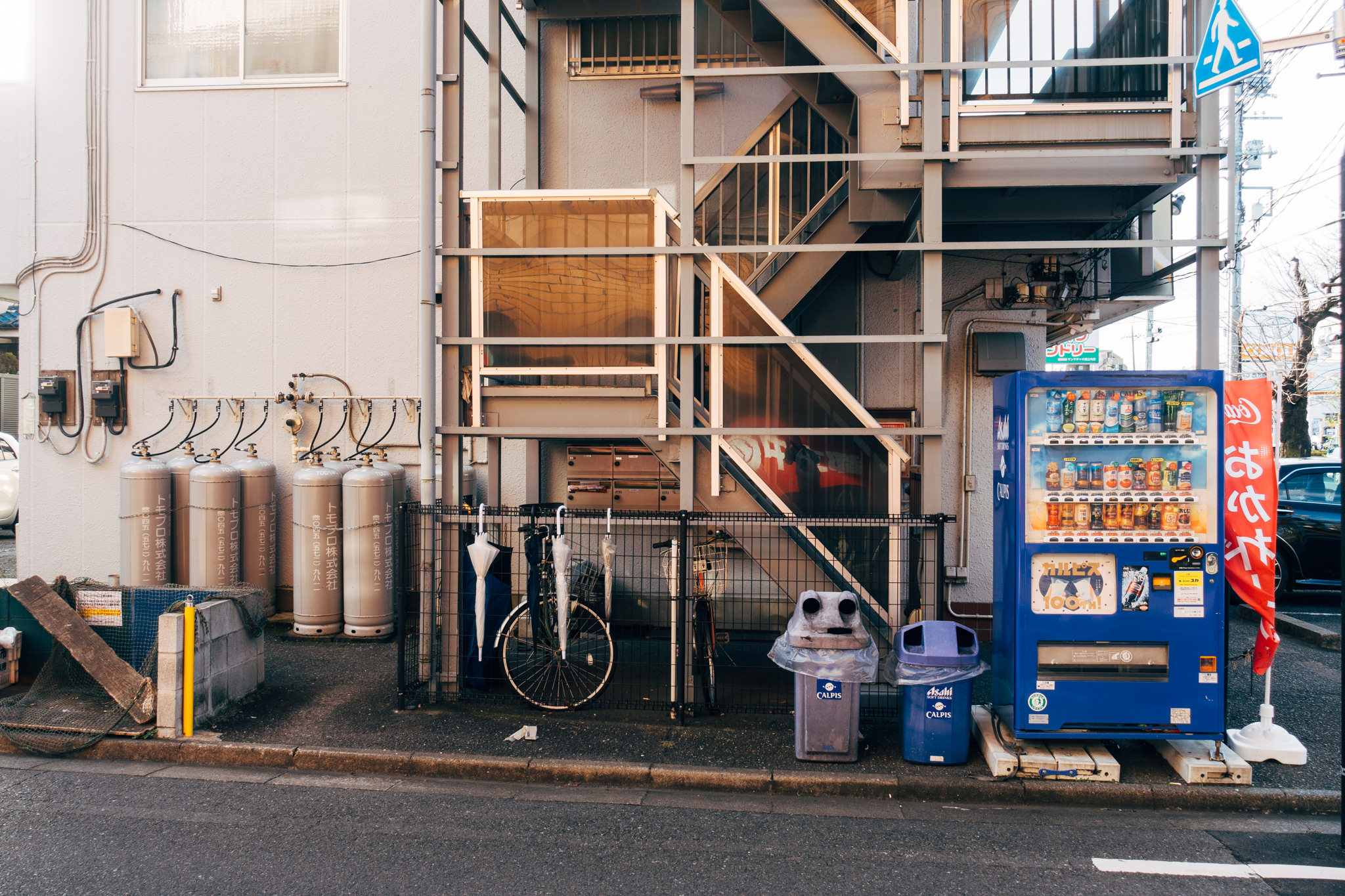 Japanese building exterior with propane tanks, a bicycle, vending machine, and trash cans.