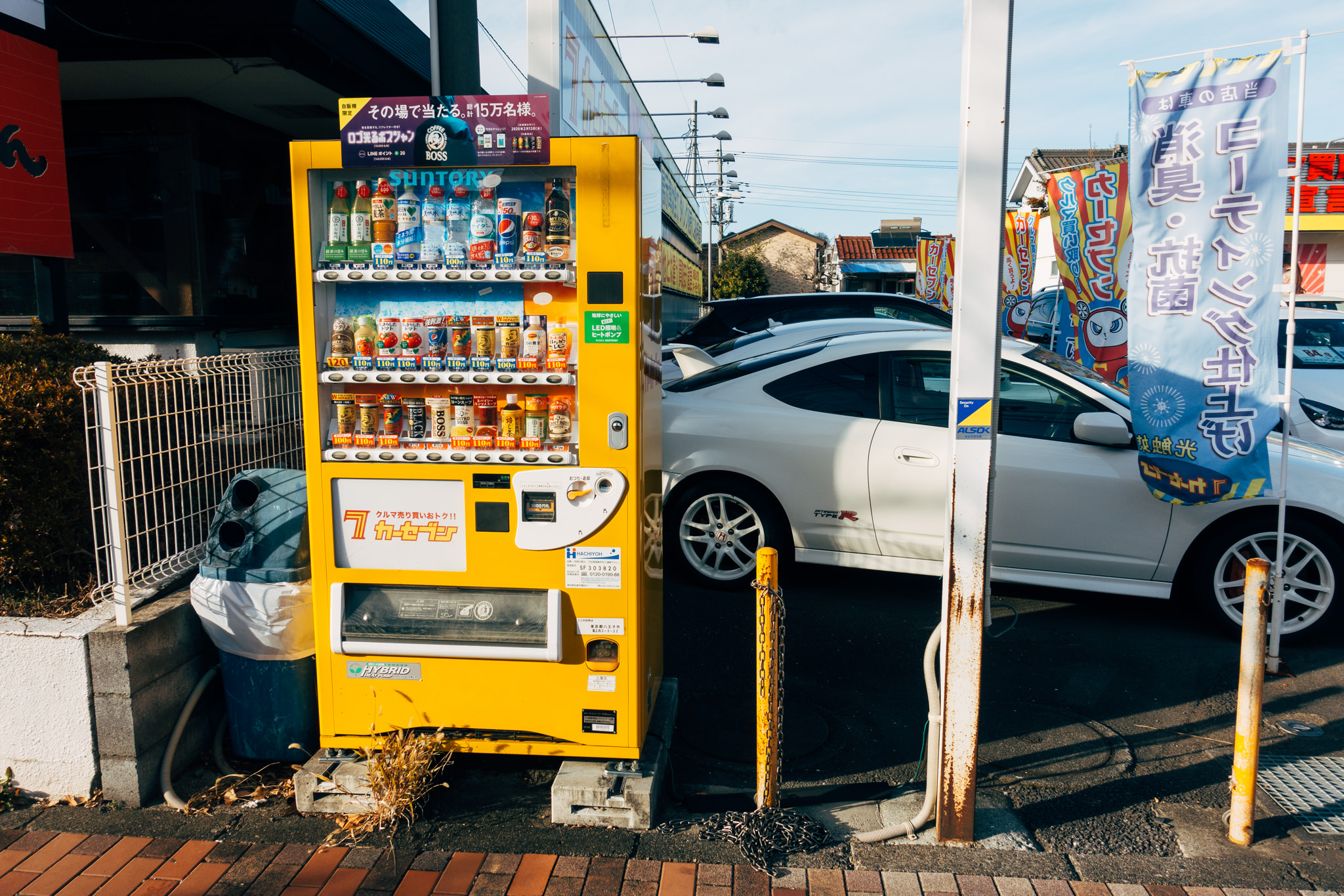 Yellow vending machine in Japan stocked with beverages, next to a white car.