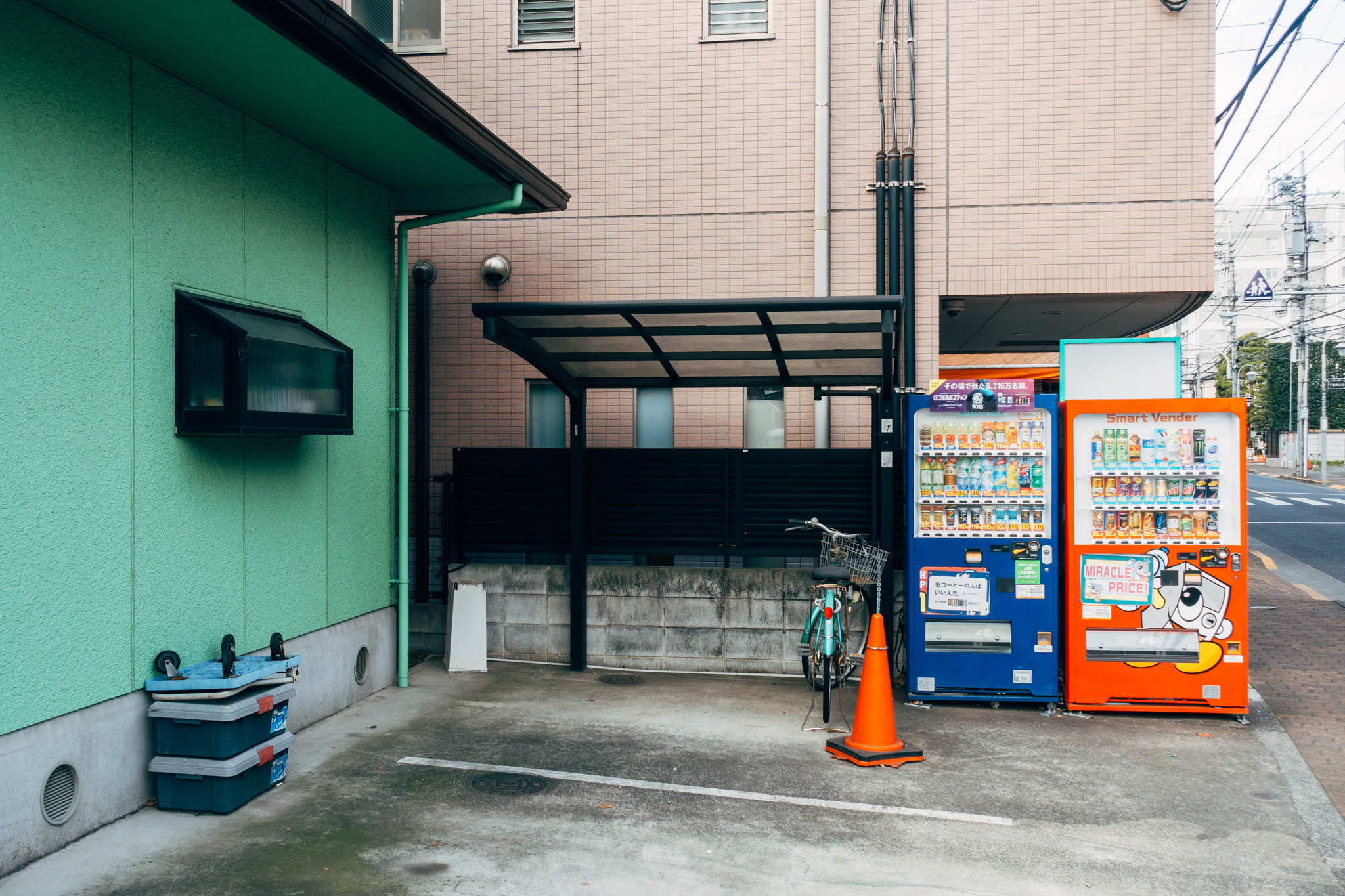 Two Japanese beverage vending machines, one blue, one orange, under a covered area; a bicycle is parked nearby.