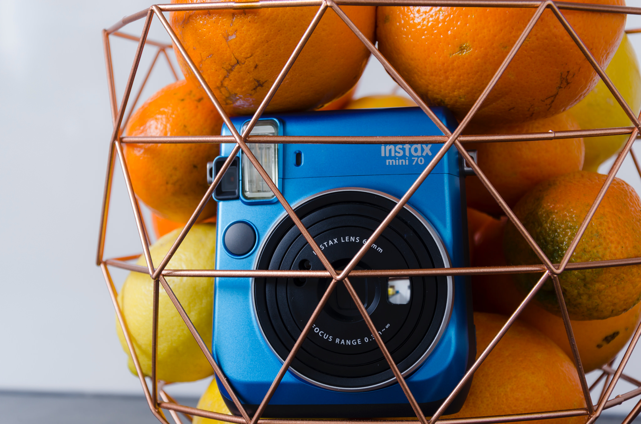 Blue Instax Mini 70 instant camera in a copper wire fruit bowl with oranges and lemons.
