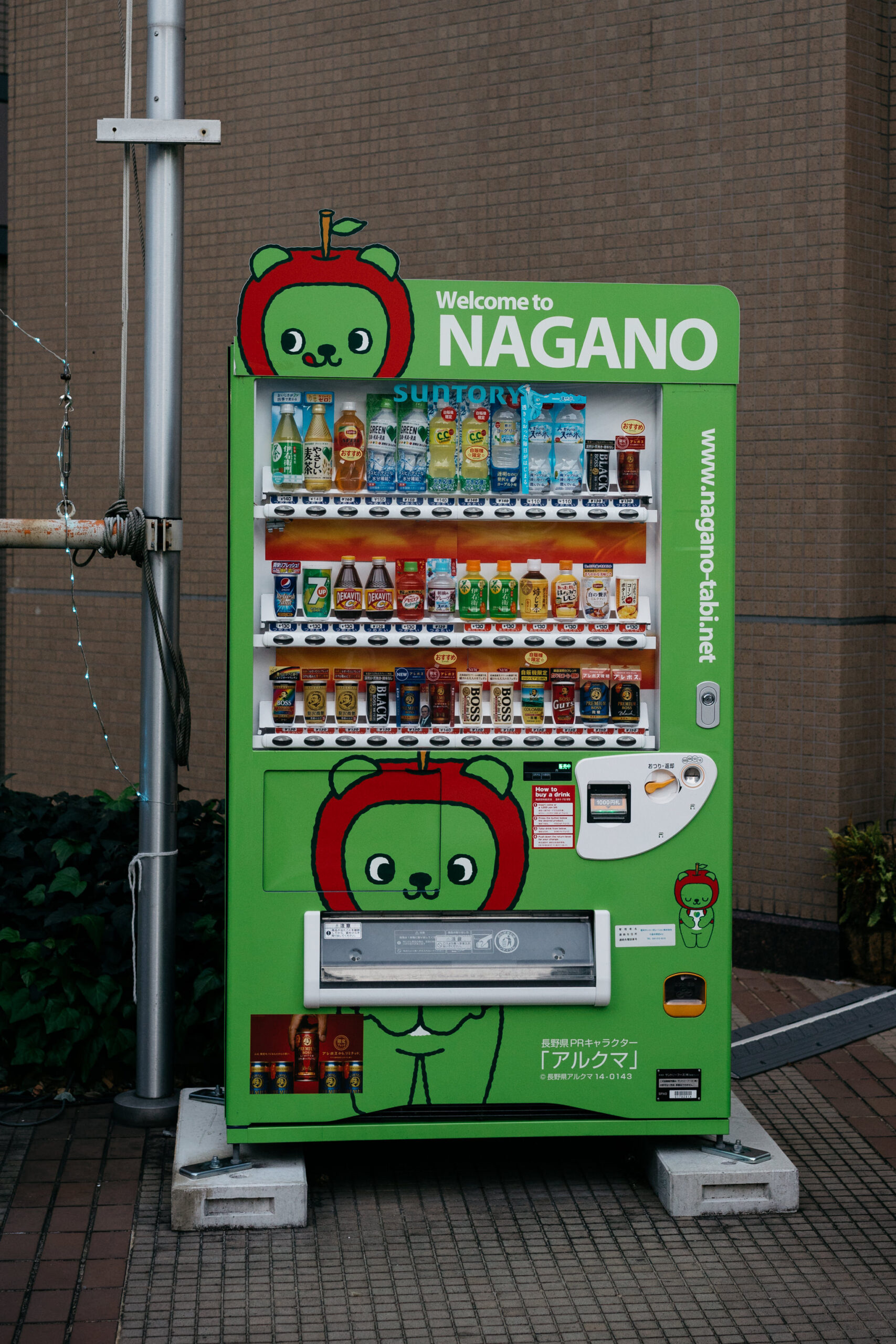Green Nagano vending machine with cartoon apple character, stocked with Suntory drinks.
