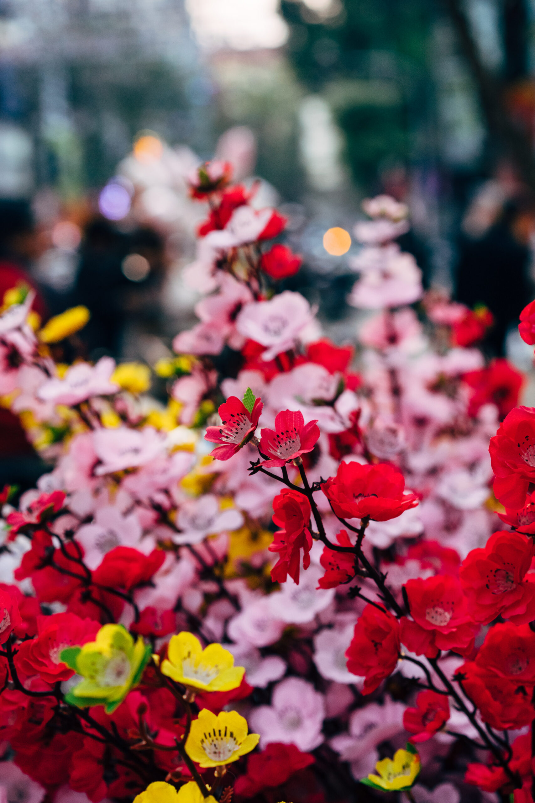 Close-up of artificial flowers in red, pink, and yellow, blurred city background.