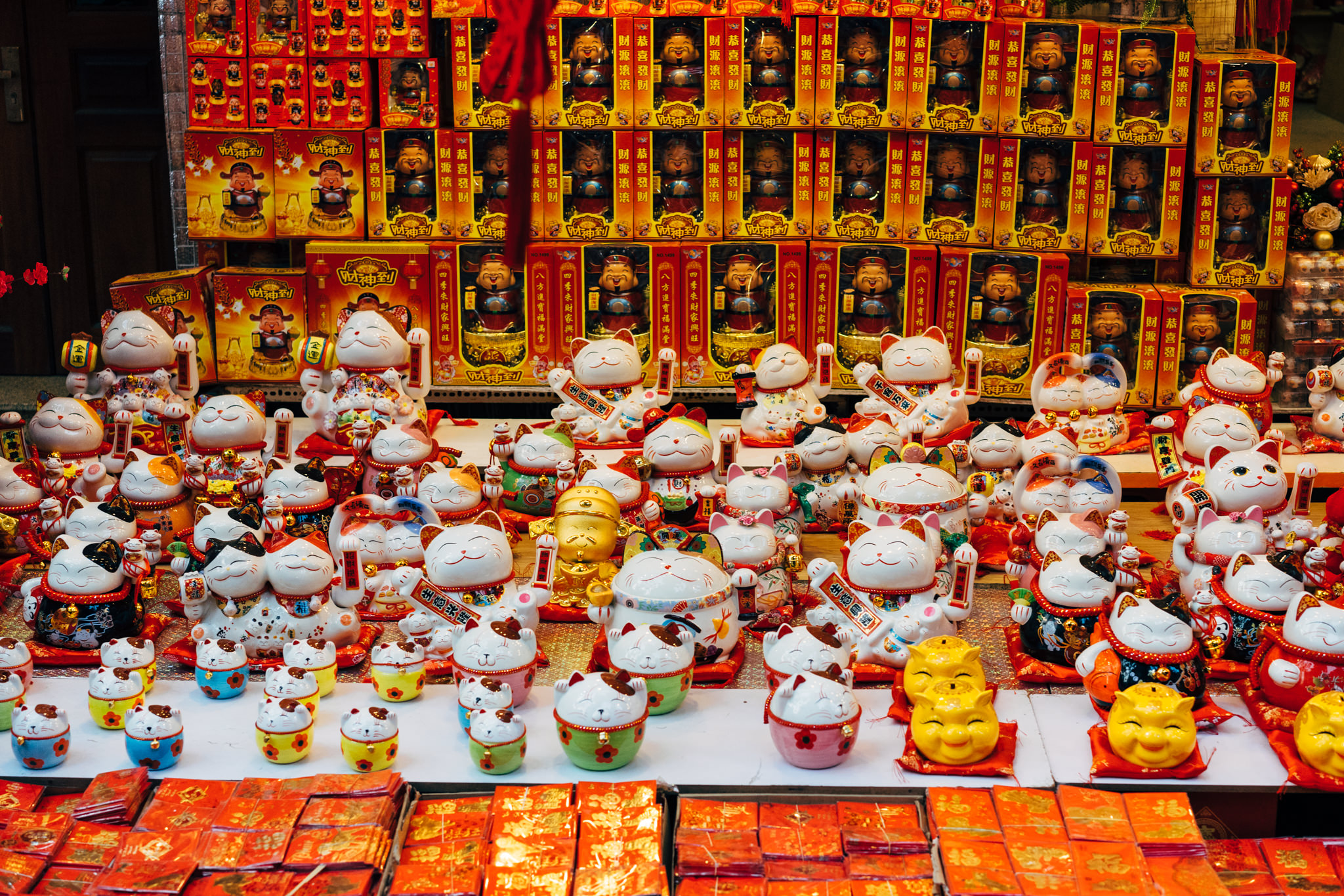 Many Maneki-neko (beckoning cat) figurines displayed on a shelf with red and gold packaging.