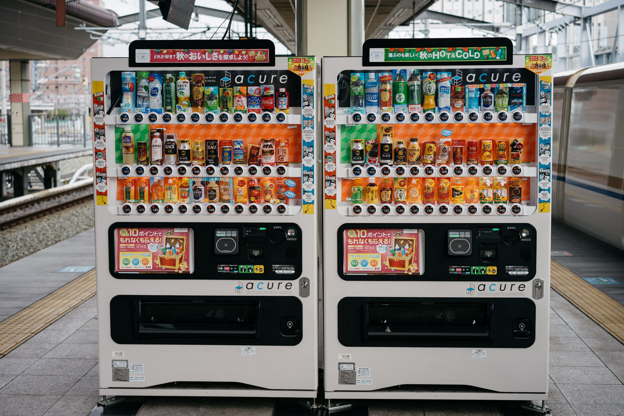 Two Acure brand vending machines stocked with various drinks at a train station.