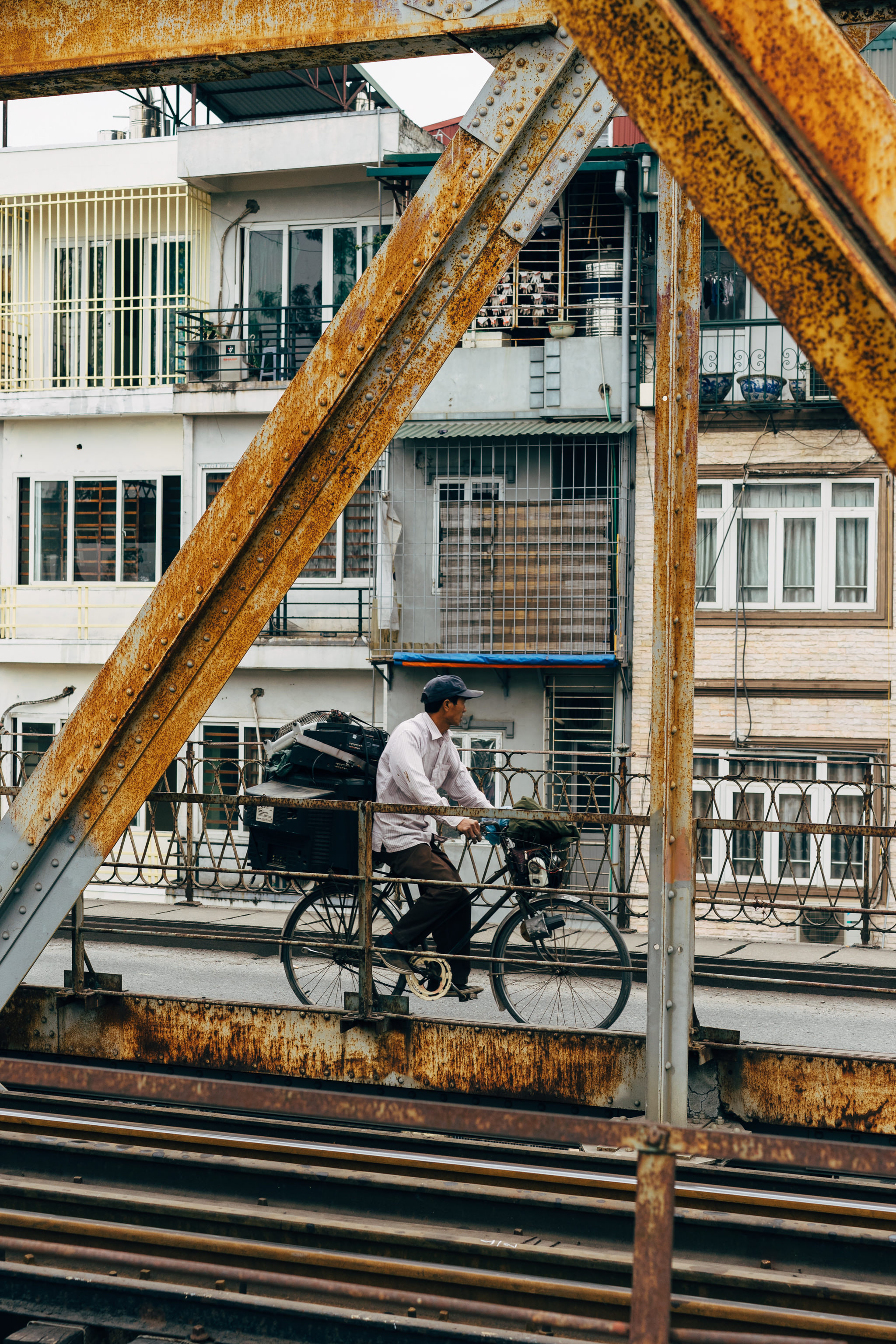 Man on bicycle carrying goods on a rusty bridge.