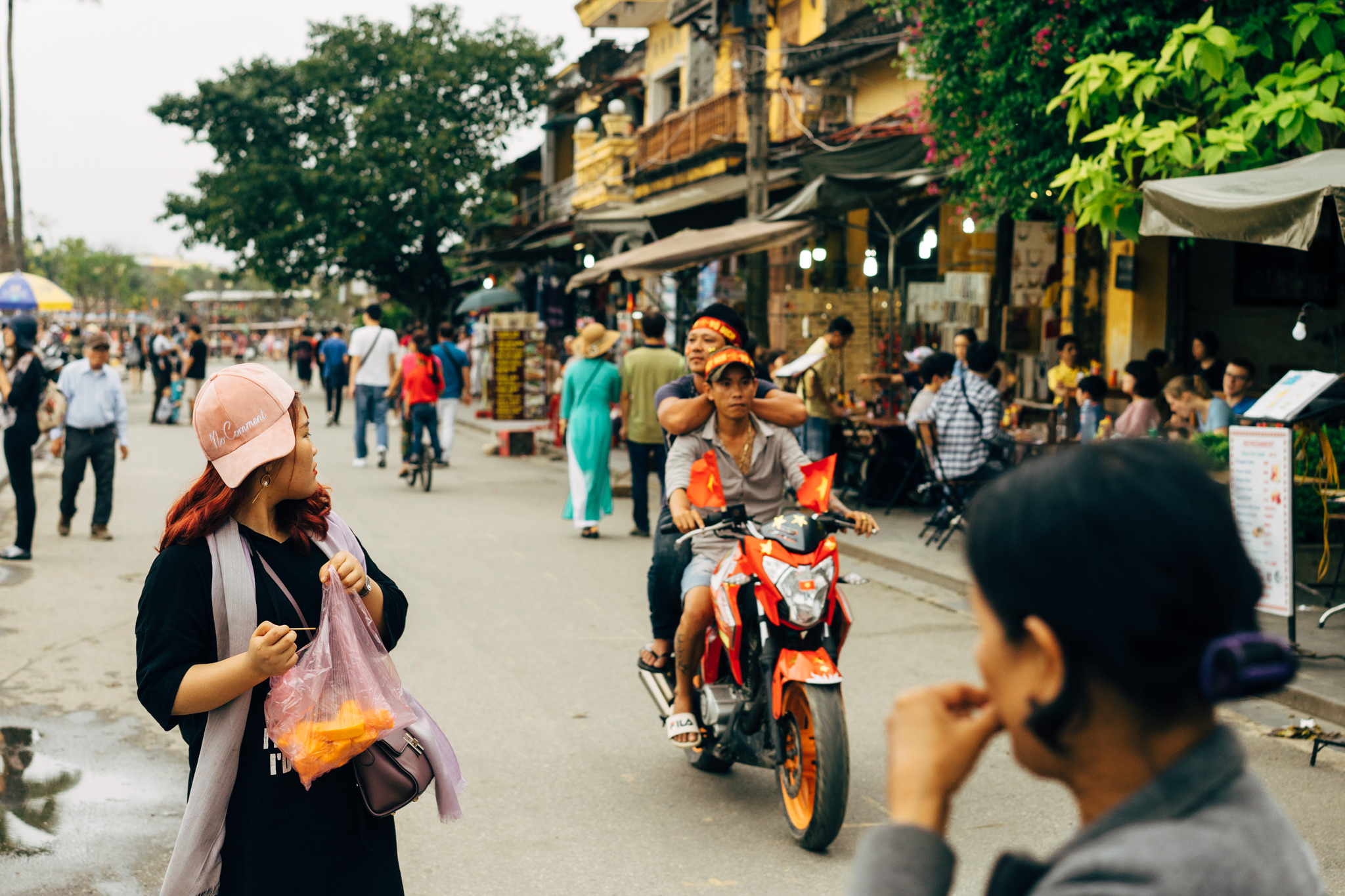 Woman in pink hat and black shirt holds plastic bag on a busy Vietnamese street, with a motorbike and many people in the background.