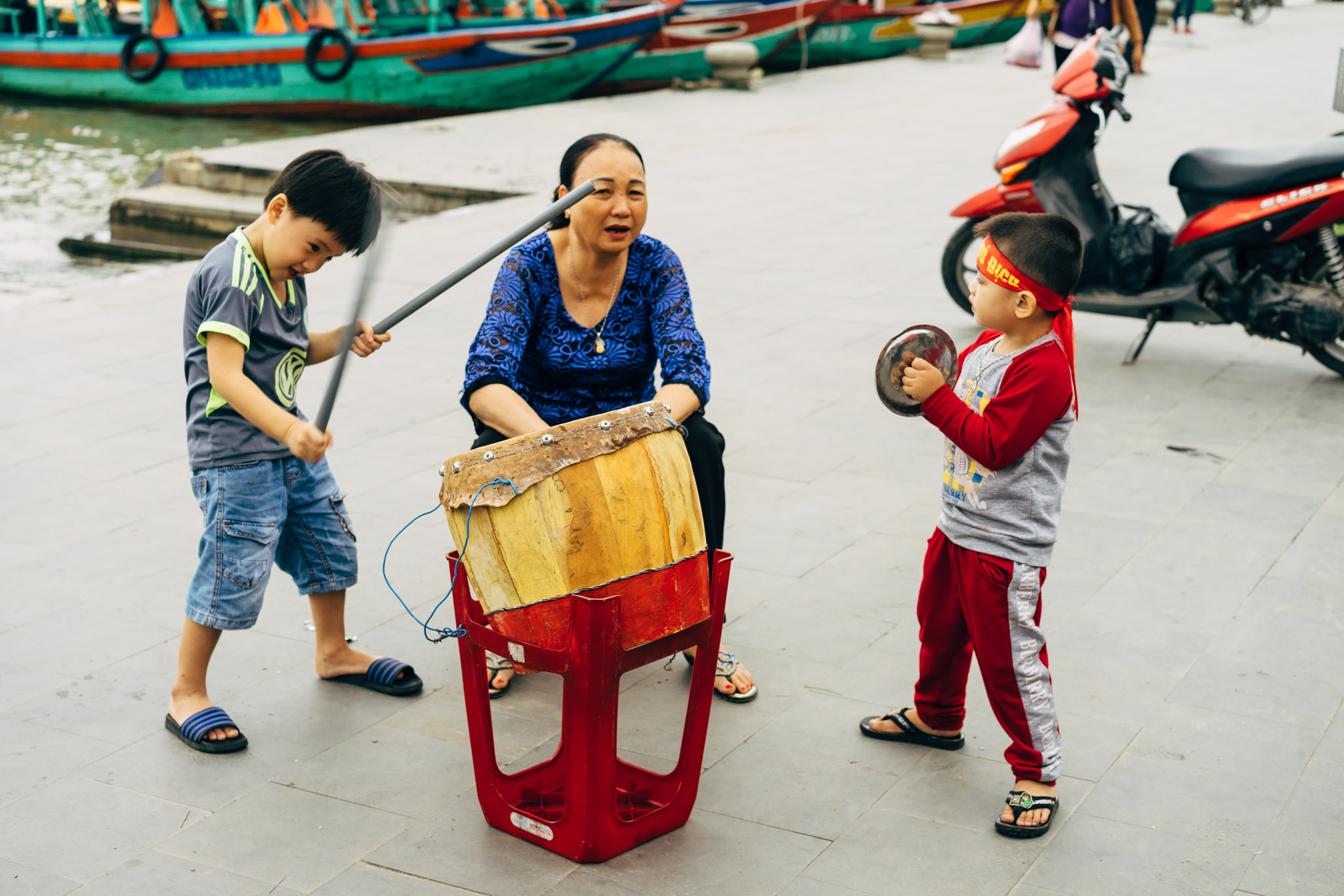 A woman and two children playing drums and cymbals outdoors.
