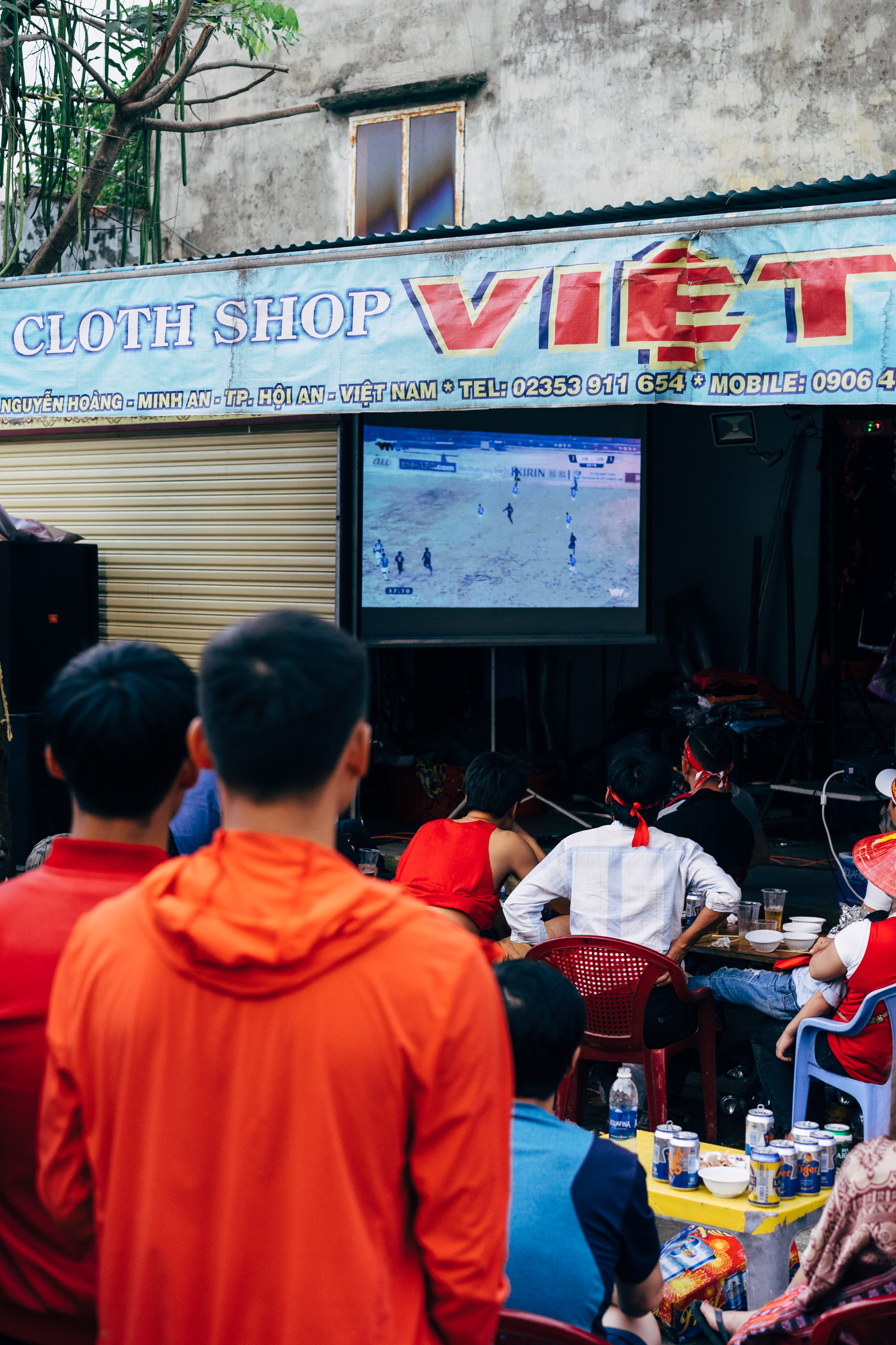 People in red shirts watch a soccer game on a large screen TV outside a cloth shop in Vietnam.