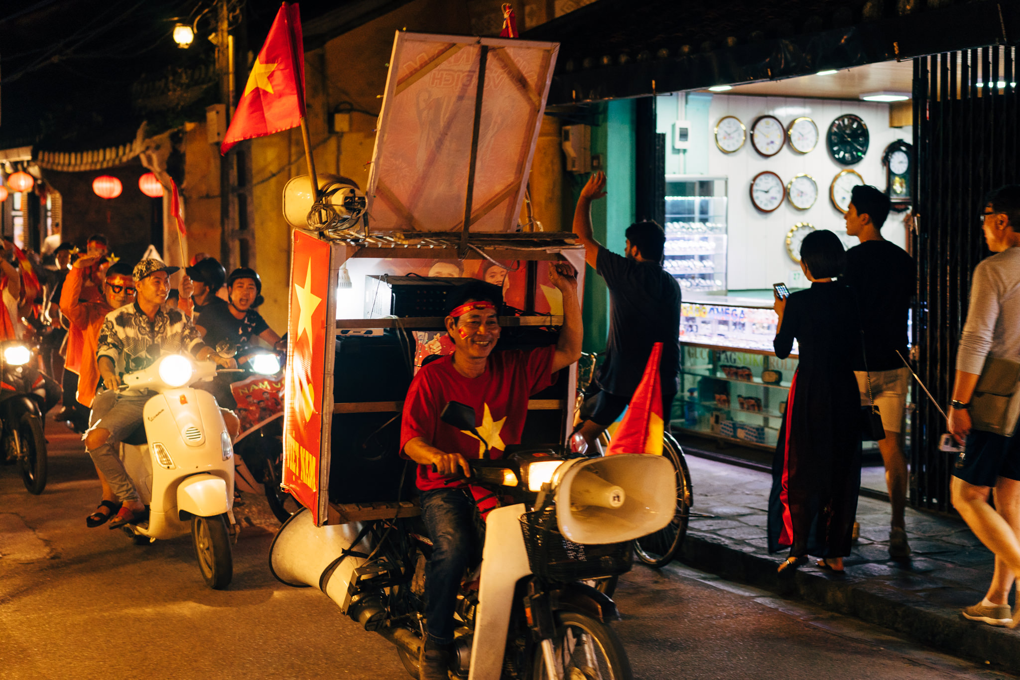 Vietnamese soccer fans celebrating in the street at night on motorbikes, decorated with flags and a sound system.