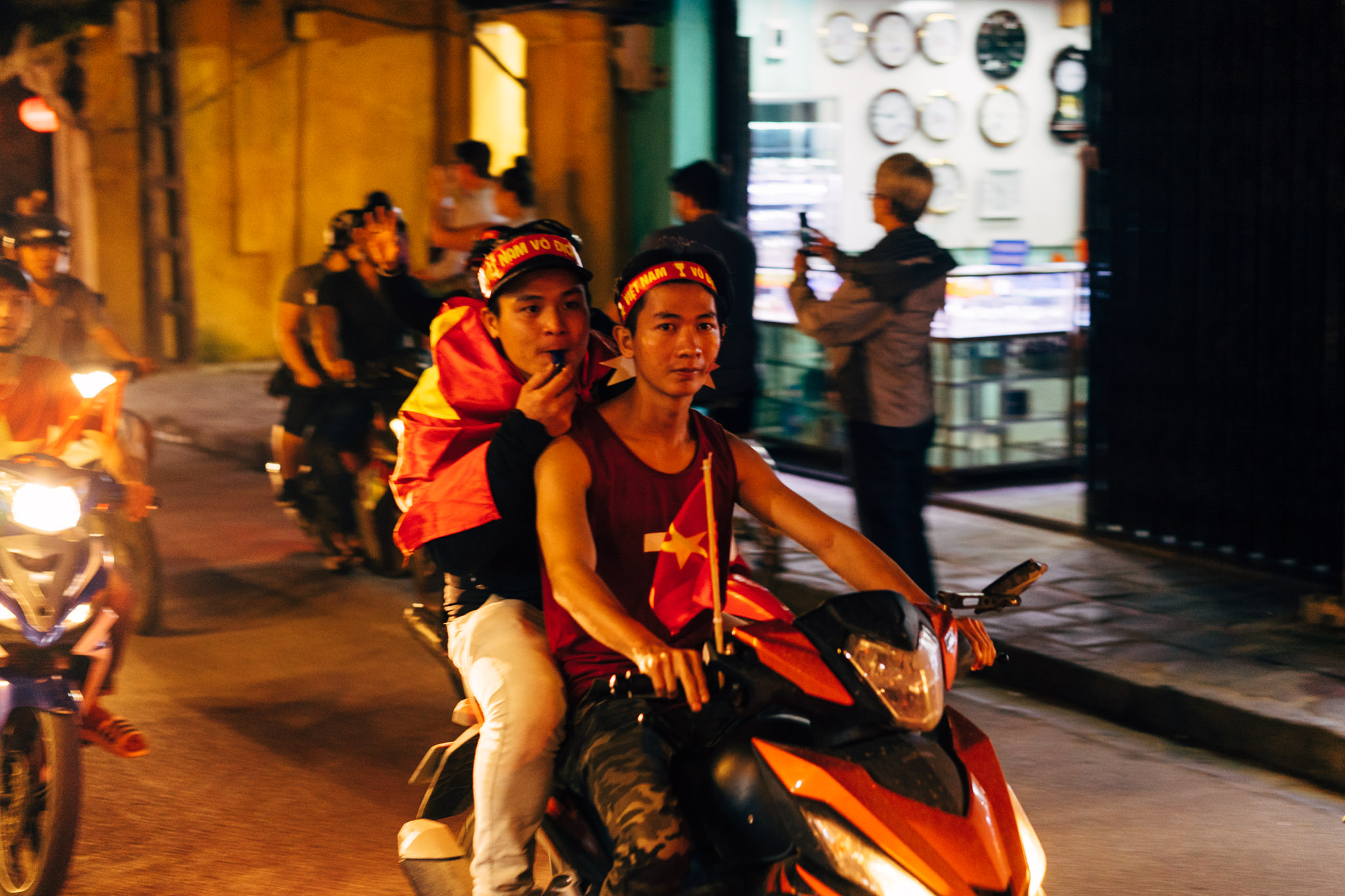 Two Vietnamese men on a motorbike at night, wearing red and yellow team colors and waving a flag.
