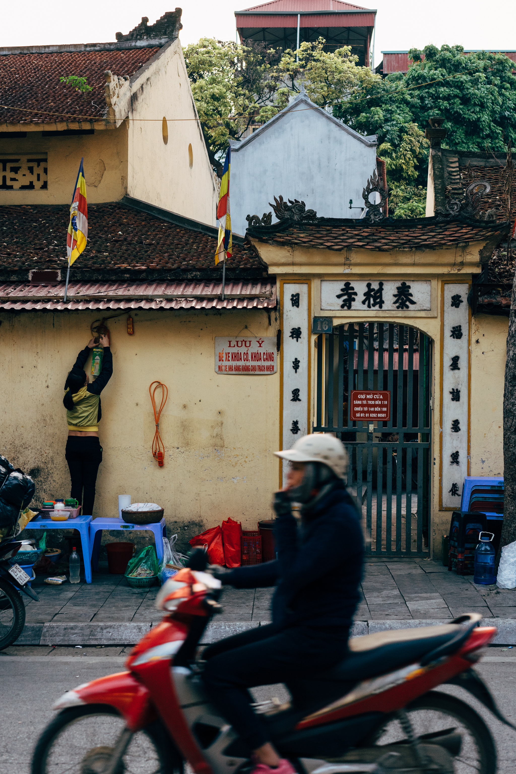 Busy Vietnamese street scene with a scooter in motion passing a person hanging a sign on a building.
