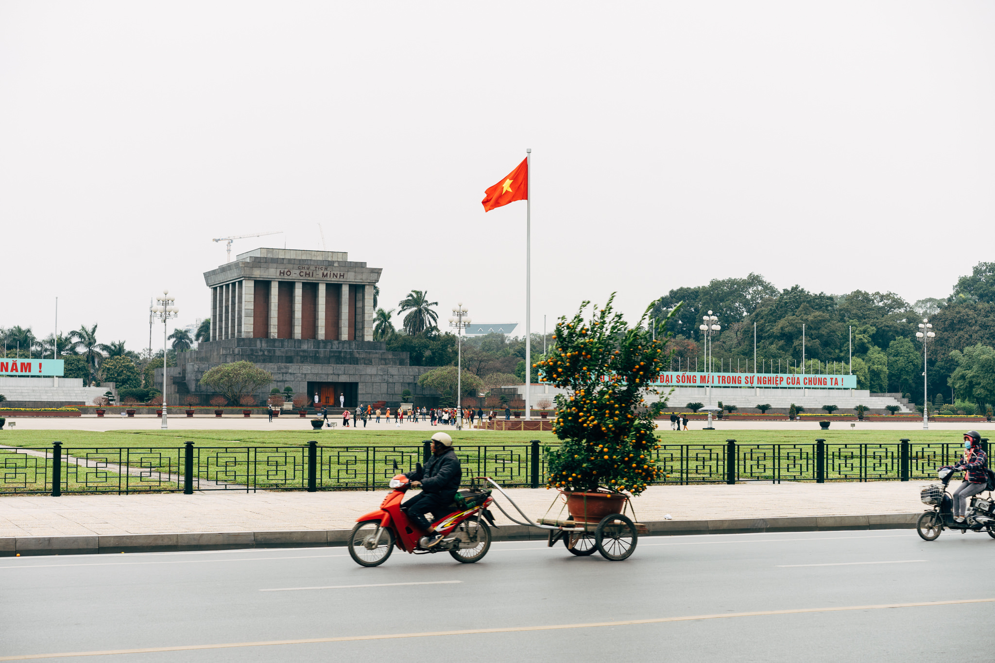 A red motorbike towing a large potted citrus tree in front of Ho Chi Minh Mausoleum in Hanoi, Vietnam.