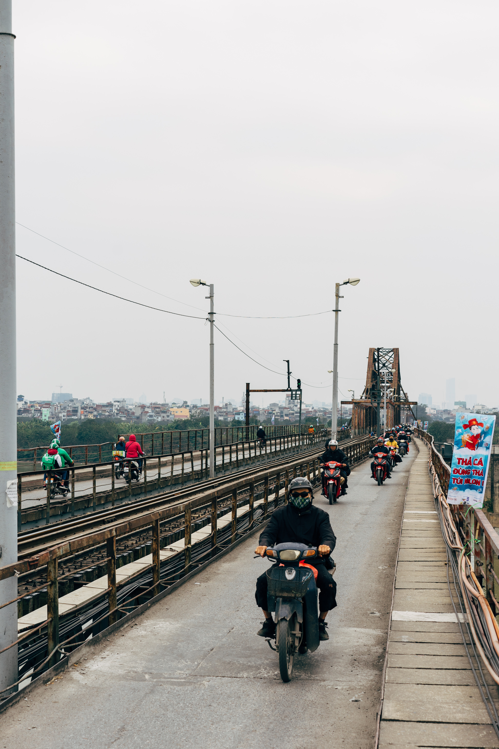 Multiple motorcyclists and scooters travel across a bridge with train tracks.