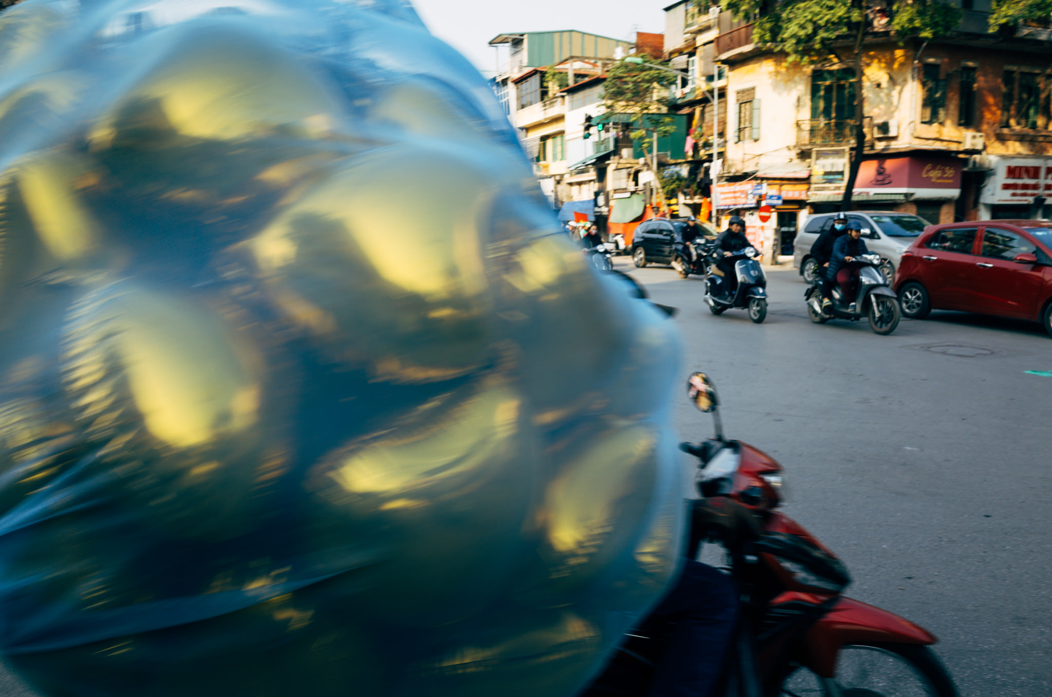 A blurred image of a person on a scooter carrying a large, plastic-wrapped bundle of yellow objects, likely balloons, amidst busy street traffic in Vietnam.