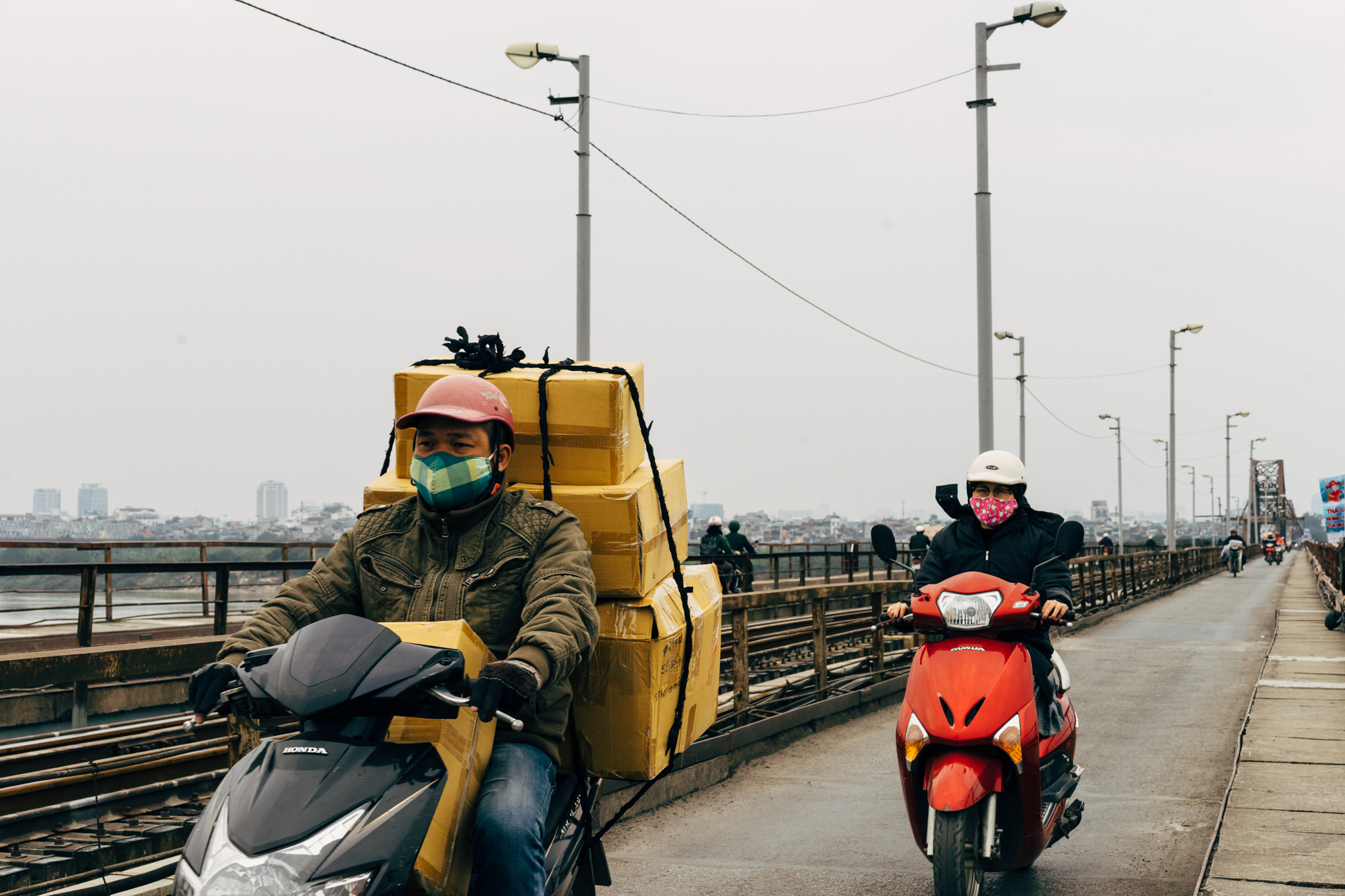 Two people wearing face masks ride scooters on a bridge; one scooter is heavily loaded with cardboard boxes.