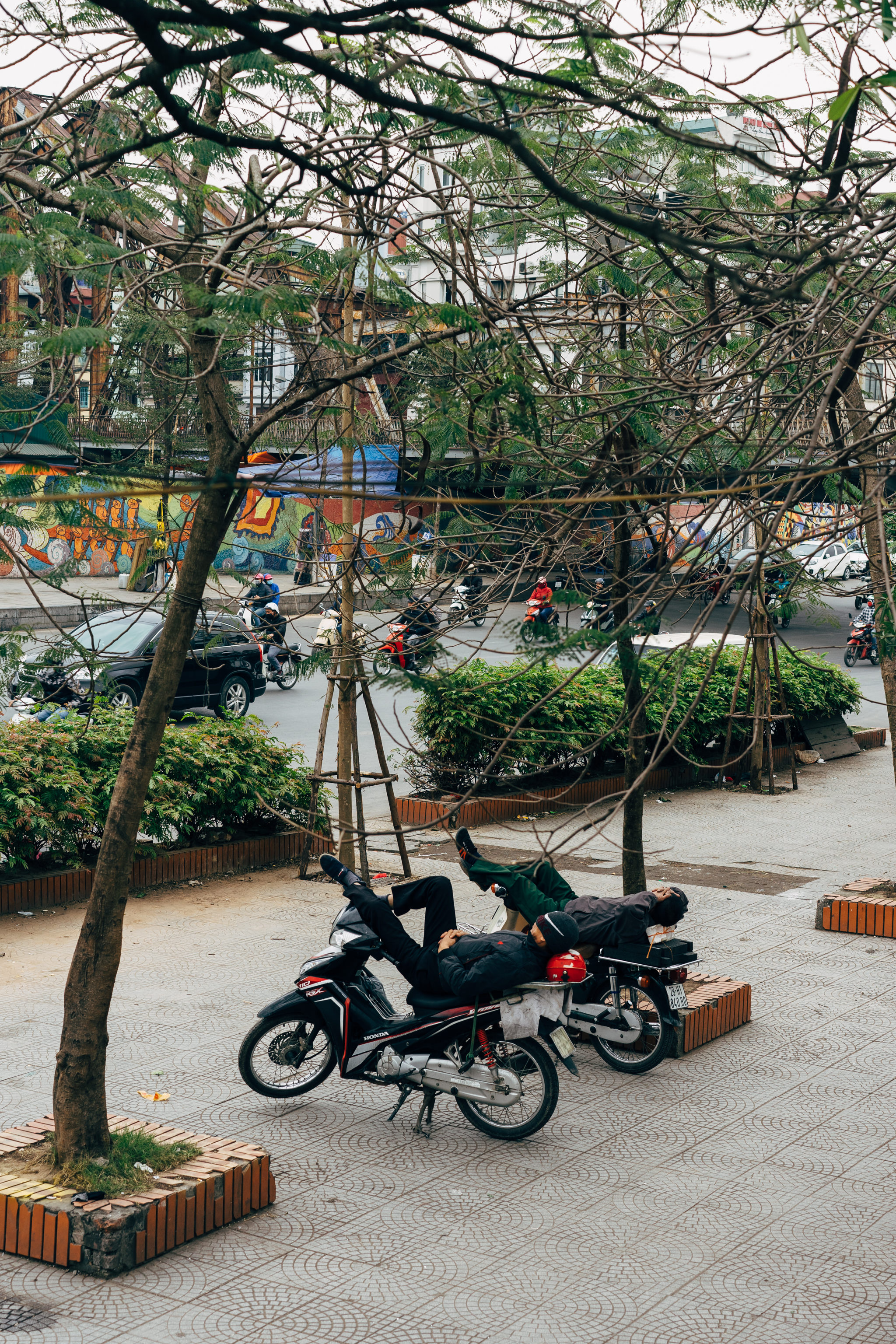 Two men sleeping on scooters parked on a sidewalk in Vietnam.